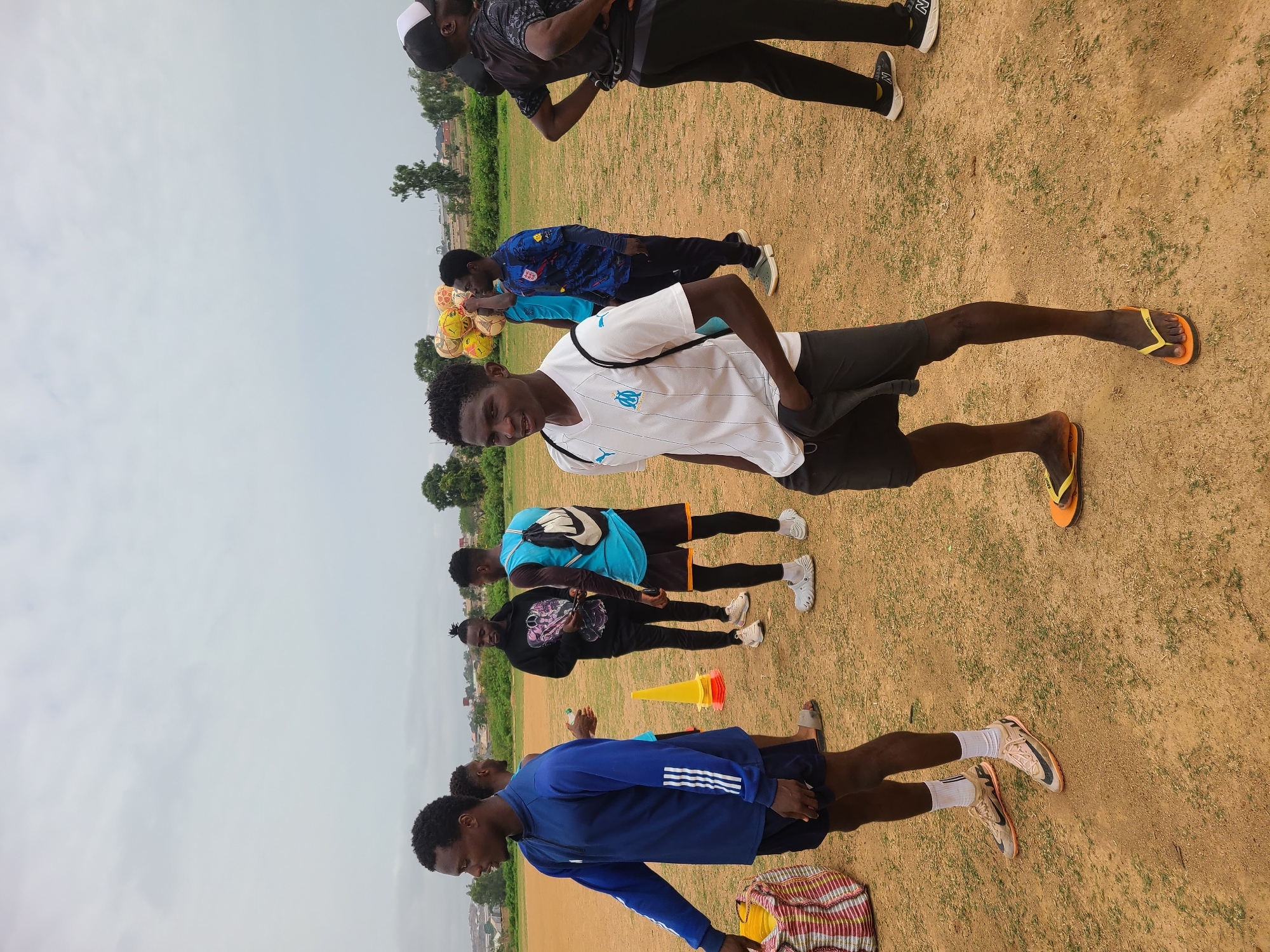 The scene is an outdoor field in Barikin Jirga Summa, Nigeria. A group of at least eight young men are present. They appear to be associated with soccer or sports, as evidenced by several soccer balls and training cones present. The environment is a grassy, dirt field, with trees and buildings visible in the background. The sky is overcast.
Several of the individuals are wearing athletic attire, including soccer jerseys and shorts. One man in the foreground is wearing a white jersey with the Marseille logo. Some are stretching or warming up. One individual is holding a bunch of soccer balls. The attire of the people suggests a warm climate.