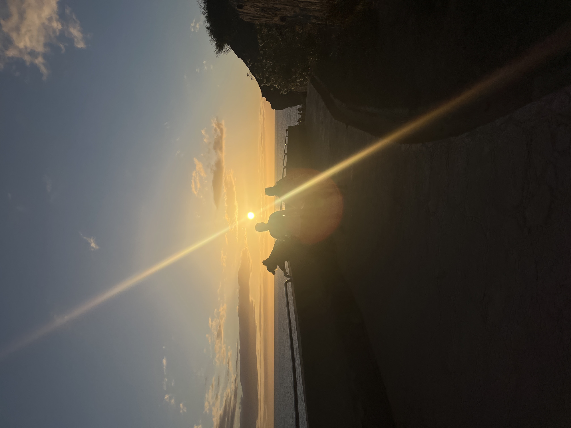 The image shows a sunset over the ocean, likely taken in Puerto Rico, Spain. A silhouette of a person or people is visible on a ledge or wall in the foreground, potentially sitting or standing. The sun's rays are prominent, causing a lens flare. The sky is a gradient of blue to orange, with some scattered clouds. A dark cliff or vegetation is visible on the right side of the frame. The perspective of the image is rotated.