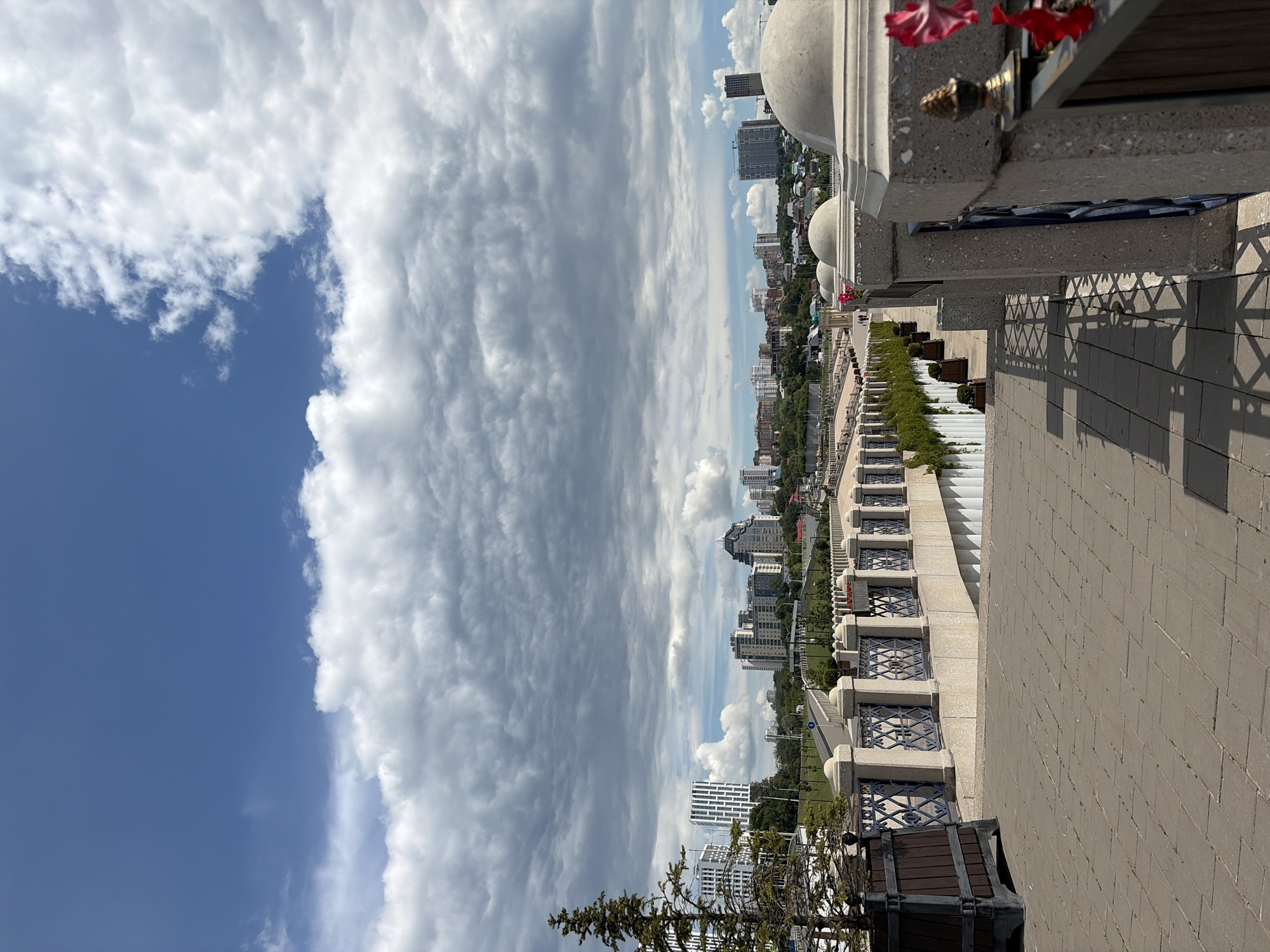 A high-angle, full shot captures a cityscape under a dramatic sky. The image is oriented vertically, with the sky occupying the left half and the city and a foreground promenade on the right.
The sky is a dynamic mix of deep blue patches and vast, turbulent clouds, ranging from bright white cumulus to darker, more ominous formations. These clouds create a sense of movement and depth.
The cityscape unfolds in the background, characterized by a dense collection of buildings with diverse architectural styles, from modern high-rises with glass facades to older, more traditional structures. Greenery is interspersed throughout the urban landscape, suggesting parks or tree-lined streets.
In the foreground, a wide, paved promenade made of light-colored bricks leads away from the viewer. To the left of the promenade, a decorative balustrade with stone pillars and ornate metalwork overlooks a terraced area. This terraced area features planters with lush green foliage and is lined with more of the decorative balustrades. A few people can be seen walking along the promenade and the terraces, indicating a public space.
The architecture of the foreground promenade includes curved, white domed structures, possibly part of a larger building or monument. Red flowers are visible in a planter near the top right corner of the image, adding a small splash of color. A dark wooden bench or seating area is positioned on the promenade.
The lighting suggests it is daytime, likely mid-morning or afternoon, given the clear blue sky patches and the shadows cast by the buildings and foreground elements. The weather appears variable, with both sunshine and cloud cover. The context provided places the scene in Ufa, Russia. No visible text is present.