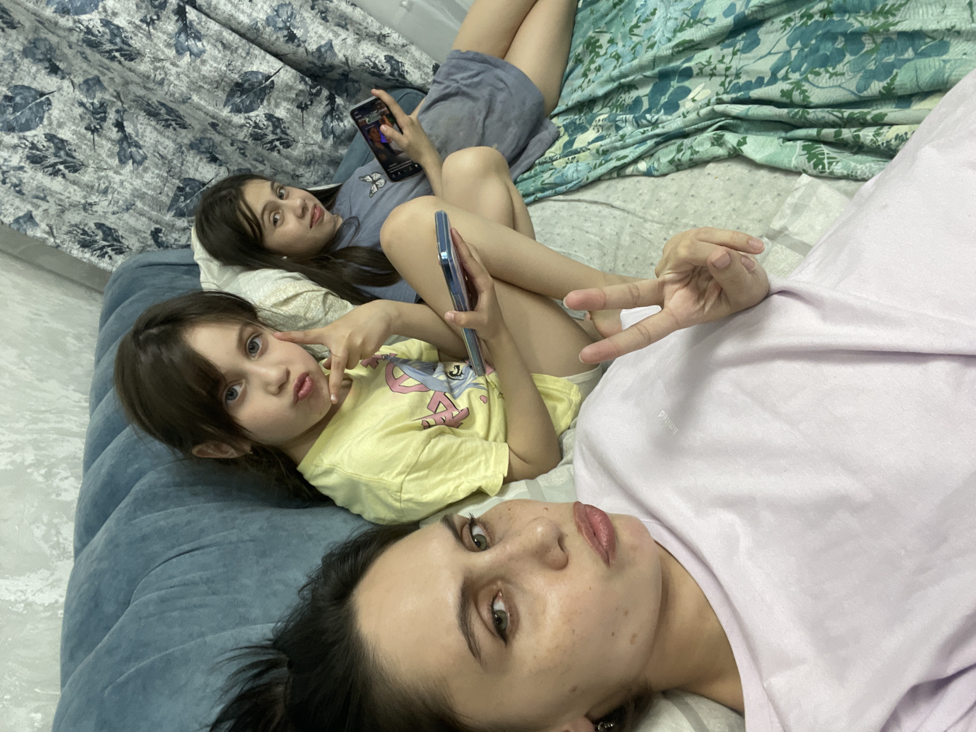 Three individuals are lying down on a bed, likely indoors. A woman, closest to the camera, has dark hair, light skin with freckles, and is wearing a lavender-colored shirt. She is looking directly at the camera. A young girl with brown hair and light skin is lying next to her, wearing a yellow t-shirt. She is holding a phone and making a "peace" sign. Further up, another girl, also with brown hair and light skin, is looking at a phone she is holding. The bed has a blue headboard and is covered with patterned sheets. The background includes curtains with different patterns, one floral and one leafy. The scene appears to be casual and informal. There is no indication of the location being Izhevsk, Russia.