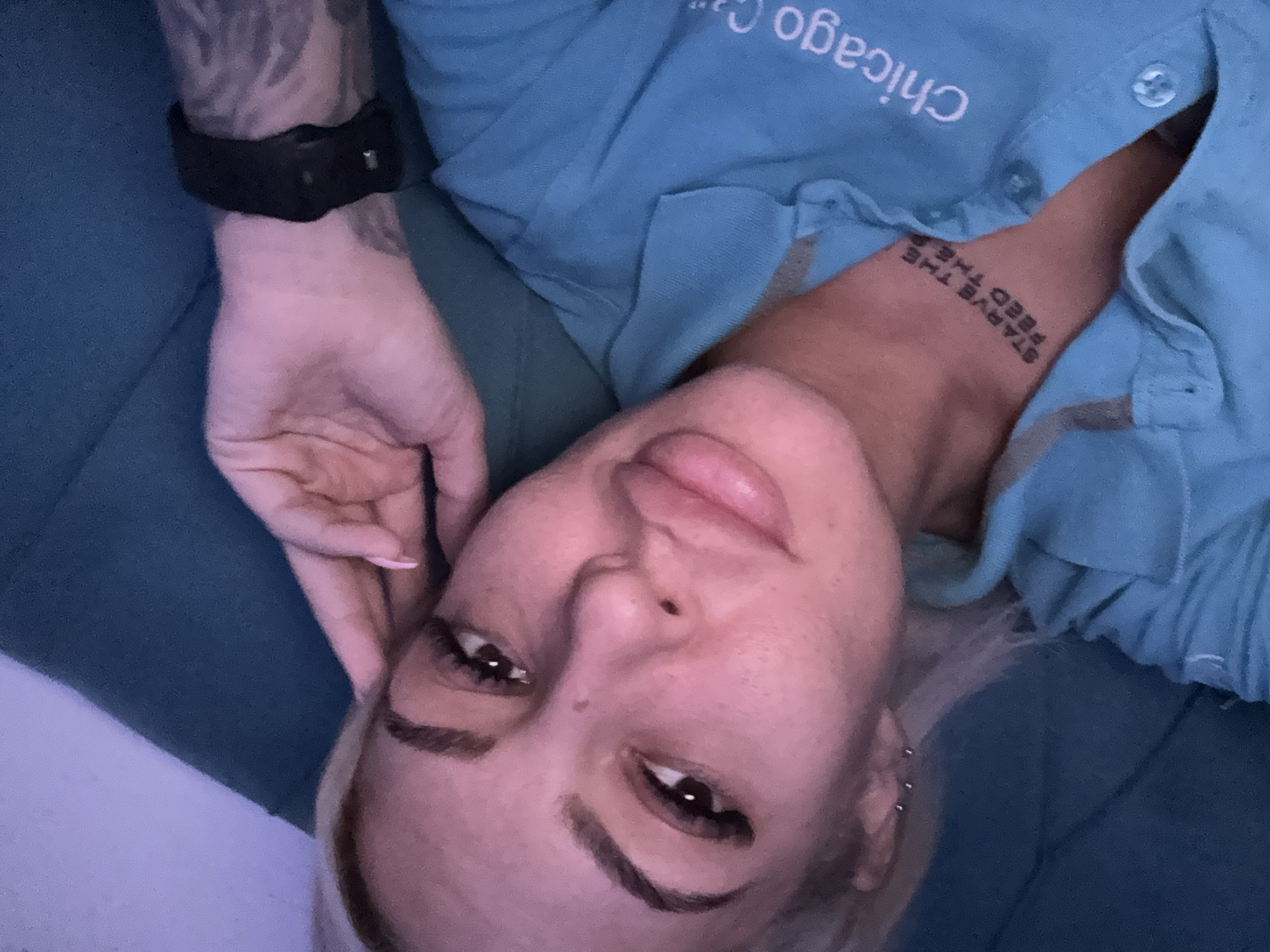 A fair-skinned individual is lying down wearing a light blue button-down shirt with the words "Chicago C" visible in white text near the collar. The person has fair hair, visible tattoos on the left forearm and neck, with a black wrist-worn device on the left wrist. They are resting their head on their left hand, and a portion of a blue cushion or padded surface is visible. The background is out of focus. The location is not depicted or identifiable from the provided image, therefore it's impossible to ascertain if it is in Plovdiv, Bulgaria.