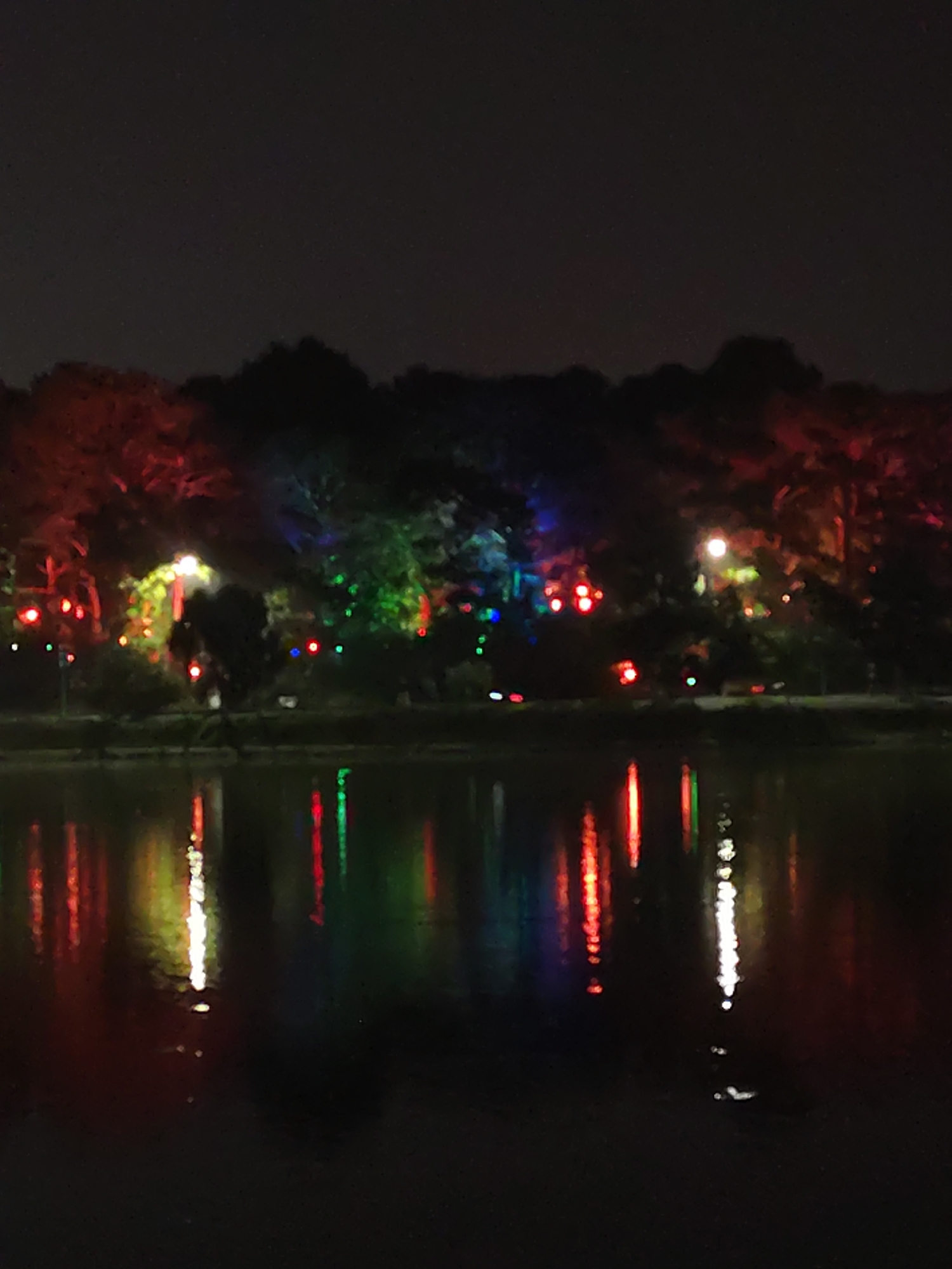 At night, colored lights are reflected in a body of water. The lights are primarily red, green, and white, and they illuminate the trees and structures along the water's edge. The image is focused on the reflected lights, with the dark water filling the foreground. The location is potentially Da Lat, Vietnam, based on visual cues. No people are visible.