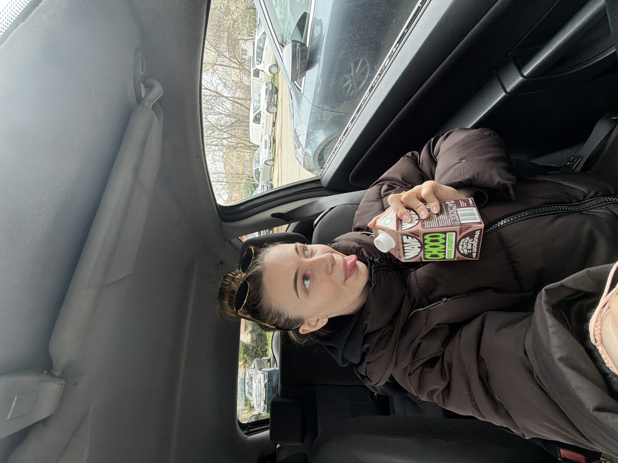 A young woman is seated in the back of a car. She's wearing a brown puffer jacket and has her hair in a bun. Sunglasses are perched on top of her head. She is holding a carton of Wave Choco chocolate milk. Her tongue is slightly sticking out. The car's interior is gray. Outside the window, other vehicles are parked along a street. Based on the visual style and the presence of cyrillic characters on the chocolate milk container, the location may be Plovdiv, Bulgaria.