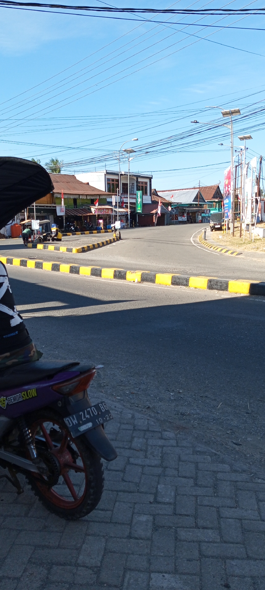 The image shows a street intersection in Pakkanna, Indonesia. A motorcycle is parked in the foreground, with its license plate clearly visible: DW. 2470. BG, with an expiration date of 10-22. A person's leg and torso, wearing a black shirt with white text, are visible on the left side of the frame, seated on or near the motorcycle. The street is paved with asphalt and bordered by a curb with yellow and black stripes. Several buildings line the intersection, some appearing to be shops or small businesses, with awnings and banners visible. Indonesian flags are displayed on some of the buildings. A few vehicles, including a small yellow auto-rickshaw and a dark van, are present in the background. Power lines crisscross the sky above, and streetlights are mounted on poles. The scene is illuminated by sunlight, casting shadows on the road. The environment appears to be a moderately developed urban or semi-urban area.