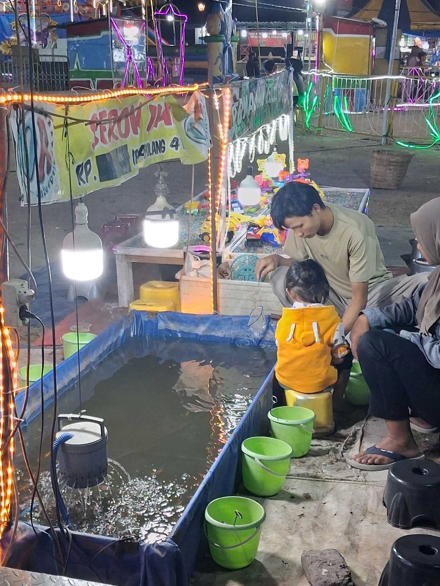 The image depicts a night scene at an outdoor amusement park or fairground, likely in Surakarta, Indonesia, characterized by colorful lights and attractions. In the foreground, a temporary pool filled with water is visible, with a filtration device actively circulating water, creating ripples and splashes. Surrounding the pool are several green buckets and a yellow stool. A young child, wearing an orange hooded jacket, is seated on the yellow stool, facing away from the camera. An adult male, wearing a light brown t-shirt, is crouched beside the child, appearing to interact with something near the pool, possibly a fishing game. Another adult, partially visible and wearing a hijab, is seated to the right. In the background, stalls and amusement rides with bright, decorative lights are set up. A banner with Indonesian text is prominently displayed, suggesting a local event. The overall atmosphere suggests a lively and family-friendly setting.