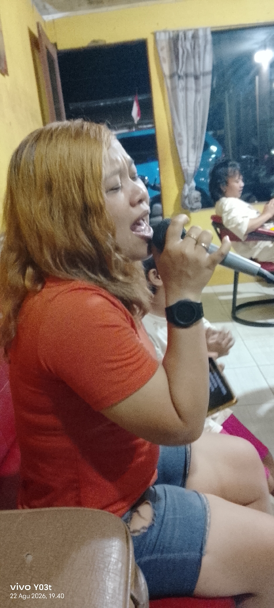 The image depicts a woman in the foreground singing into a microphone. She has light brown hair and is wearing an orange t-shirt and denim shorts. She is holding the microphone with her right hand, and her left hand is resting on her lap. Her eyes are closed, and her mouth is open as if she is singing loudly. On her left wrist, she is wearing a black watch.
In the background, another person, a woman with dark, curly hair, is sitting in a chair and appears to be looking at something in her hands, possibly a phone or a tablet. She is wearing a light-colored shirt. The environment appears to be indoors, with a yellow wall behind the main subject. A curtain is visible on the right side of the image. The lighting suggests it is evening or night.
The timestamp at the bottom of the image indicates it was taken on "22 Agu 2026, 19.40" and captured by a "vivo Y03t" device. The date format suggests the location is Indonesia, and the prompt specifies Dawidawi, Indonesia.