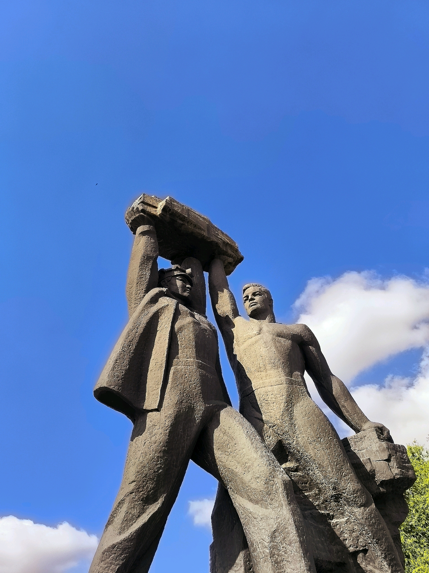 This low-angle, full-body shot captures a monumental stone sculpture of two men lifting a large slab above their heads. The sculpture is positioned outdoors against a vibrant blue sky with scattered white clouds. The figure on the left wears what appears to be a uniform with a cap, while the figure on the right is bare-chested and muscular. Both figures are depicted with serious, determined expressions. The sculpture exhibits a rough, textured finish, common in Soviet-era art. The overall impression is one of strength, labor, and unity. The sculpture is located in front of the Qarağandı City Administration building in Kazakhstan.