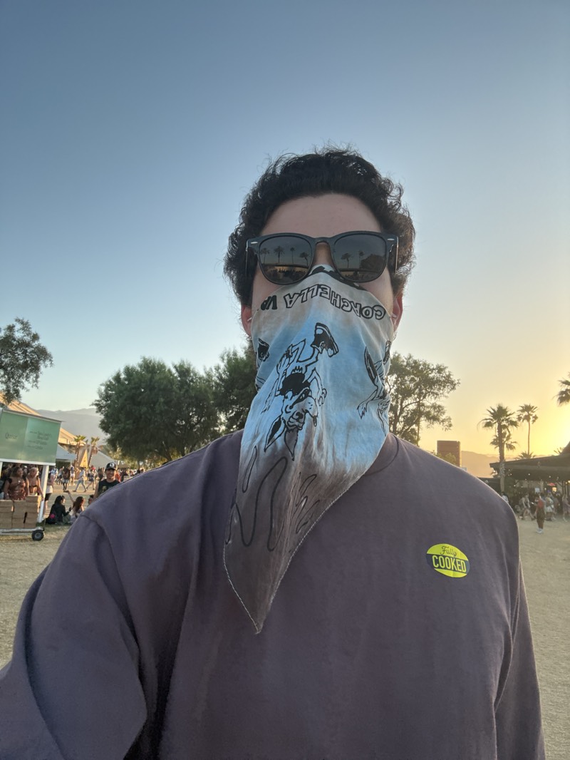 A man stands outside at the Coachella Valley Music and Arts Festival in Indio, California, United States. He wears dark sunglasses, a light blue patterned bandana covering his nose and mouth, and a brown long-sleeved shirt with a yellow "Fully Cooked" logo. Trees, people, and a setting sun fill the background.