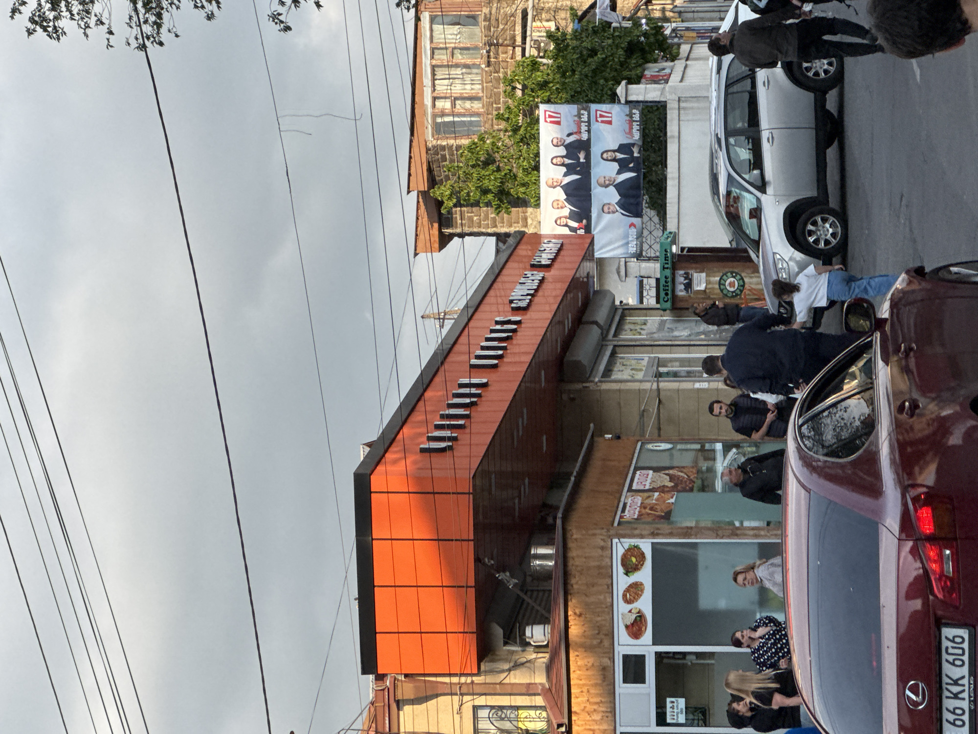 The image shows a street scene in Yerevan, Armenia. A two-story building with an orange facade dominates the foreground. The name of the business "SELVAGESTA CAFE" is displayed on the upper portion of the facade. The first floor has a glass storefront. People are standing and walking in front of the cafe. The rear of a red Lexus car is in the lower right corner, with the license plate number "66 KK 606." A silver SUV is parked on the street behind the red car. A political poster hangs above the cafe. Overhead are many overhead power lines.