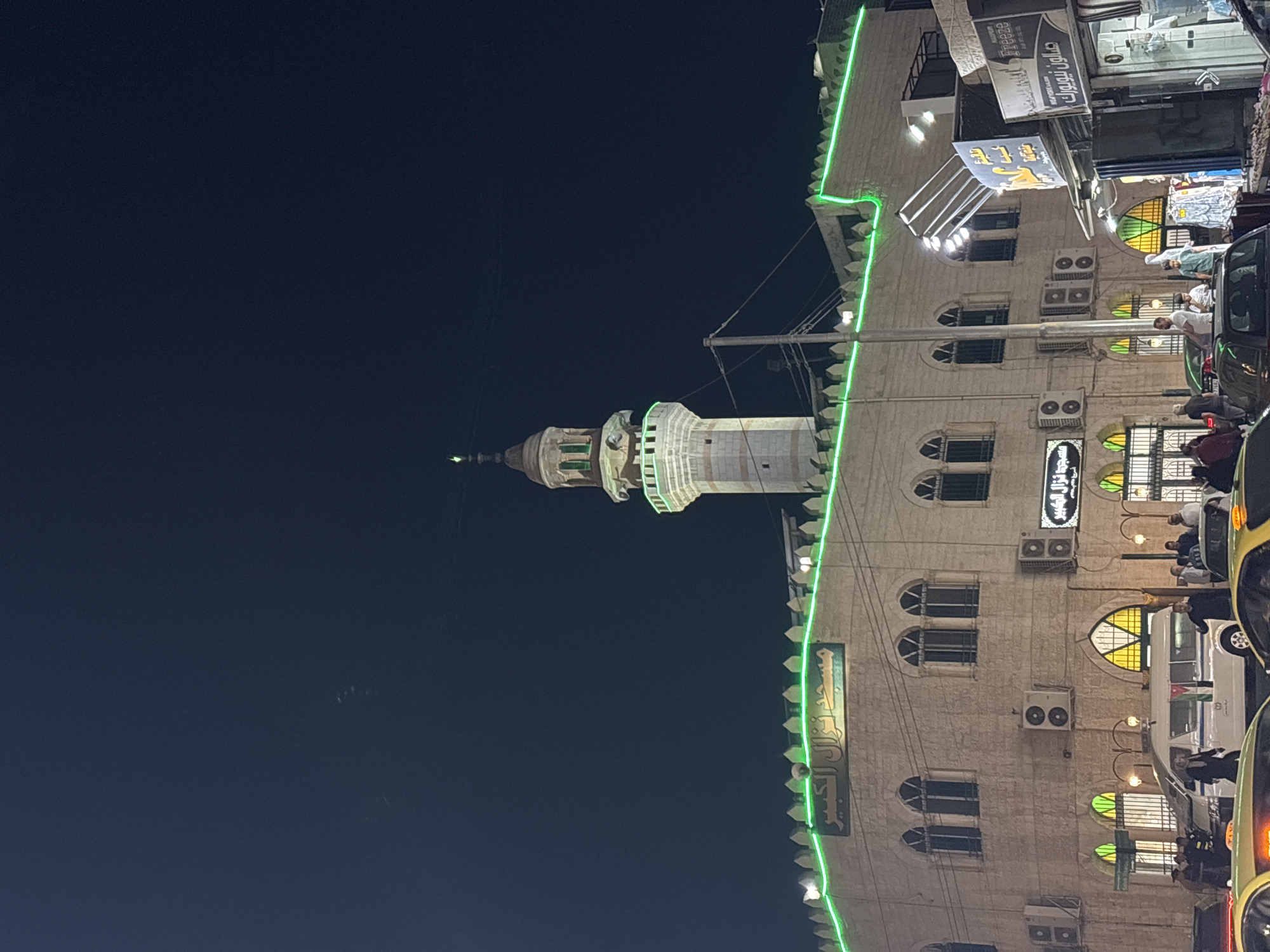 The image depicts a nighttime view of the "Nuzal Al Kabir Mosque" in Amman, Jordan. The mosque's facade is illuminated with green neon lights along its crenellated rooftop. A tall, slender minaret with a lantern at its apex rises from the right side of the mosque. Below the mosque, street level is visible with several shops. One shop sign, in Arabic script, reads "صالون نيويورك" (New York Salon). A white van with tinted windows and an Arabic inscription, possibly indicating an official vehicle with a Jordanian flag emblem, is parked in front of the mosque. Several people are seen walking on the sidewalk and around the vehicles. In the foreground, the tops of yellow taxis are visible, suggesting a busy urban street. The sky is dark, indicating it is nighttime, with a few faint lights or stars visible.