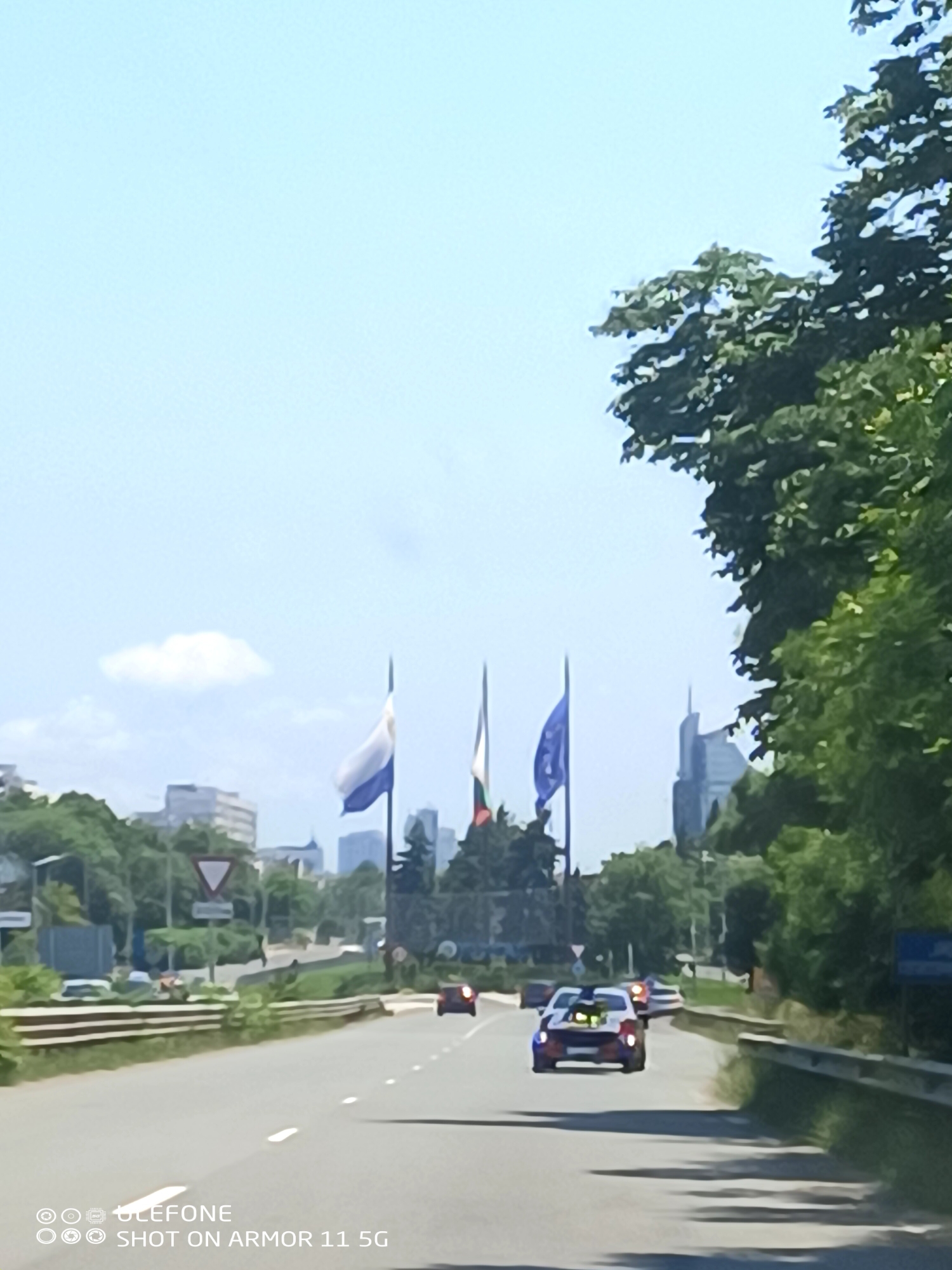 This detailed image captures a daytime scene looking down a multi-lane asphalt road leading into the city of Burgas, Bulgaria. The sky is bright light blue with a single small white cloud visible on the left.
In the immediate foreground, the asphalt road shows faded white dashed lane markings. On the right, a metal guardrail lines the road, accompanied by dense green foliage and trees. Shadows from unseen objects or trees fall across the right side of the road. The bottom left corner features a white watermark: "© ULEFONE" and "SHOT ON ARMOR 11 5G".
In the midground, several vehicles are traveling away from the viewer. Most prominently, a customized white car with blue and red accents, featuring an exposed engine or intake system protruding from its hood, occupies the middle lane. Further ahead in the right lane, a red car is visible. Other indistinct vehicles can be seen in the distance. On the left side of the road, more green trees line the area, and a "Yield" road sign (an inverted white triangle with a red border) is clearly visible. Buildings are partially obscured by the trees on the left.
The background reveals the city skyline of Burgas. Three tall flagpoles stand prominently. From left to right, the flags displayed are: a white and blue flag (its specific design is unclear), the national flag of Bulgaria (horizontal stripes of white, green, and red), and a predominantly blue flag (likely the flag of the European Union). Behind the flags and trees on the far right, a distinctive modern skyscraper with a unique angular top is visible, identifiable as the Burgas Free University building, a prominent landmark in Burgas. More city buildings are partially visible through the trees and in the distance. No people are visible in the scene.