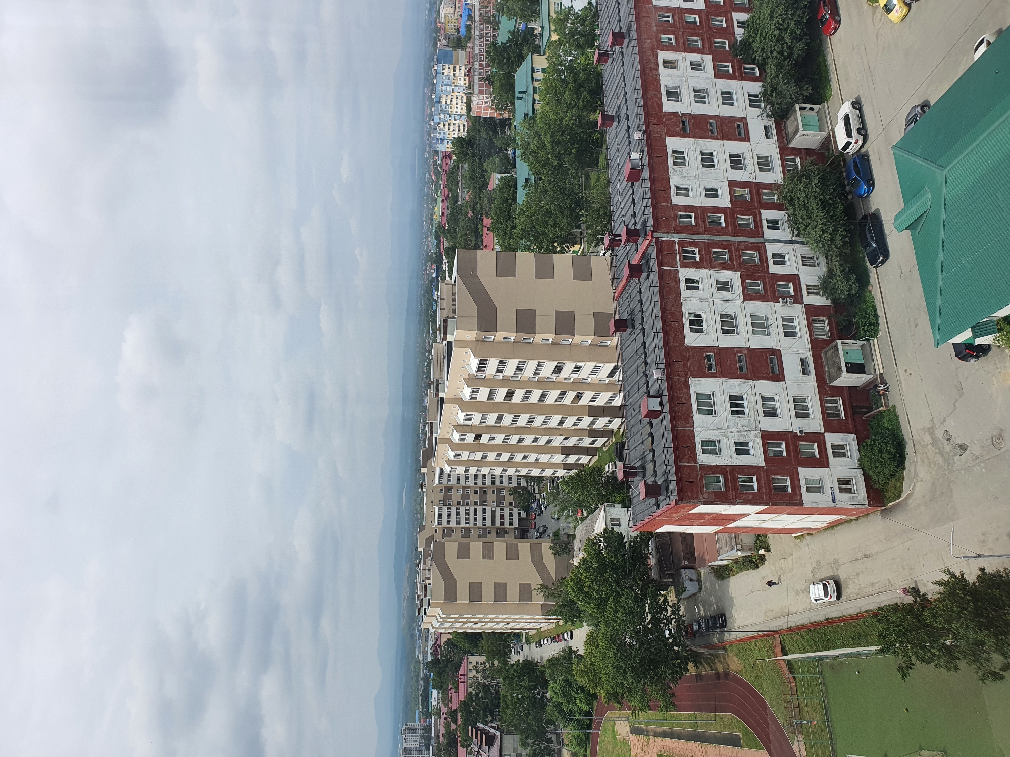 The image shows an aerial view of an urban residential area in Yuzhno-Sakhalinsk, Russia, under a cloudy sky. In the foreground, a predominantly red and white apartment building with multiple windows and a dark, multi-pitched roof with numerous ventilation pipes is visible. Several cars, including a white SUV, a black sedan, a blue sedan, and a red sedan, are parked along the street in front of this building. A portion of a green-roofed structure is also present in the lower right corner.
Further back, a row of tall, beige apartment buildings with repeating window patterns and cream-colored facades are prominent. These buildings are situated along a street lined with parked cars. Green trees are interspersed throughout the scene, particularly around and between the buildings. To the left, a sports field with a red running track is partially visible.
In the background, more residential buildings of varying colors and heights, including a blue and white structure, are visible, along with a dense cluster of trees. Distant, hazy mountains form the horizon under the overcast sky. The overall impression is that of a typical multi-story residential district.