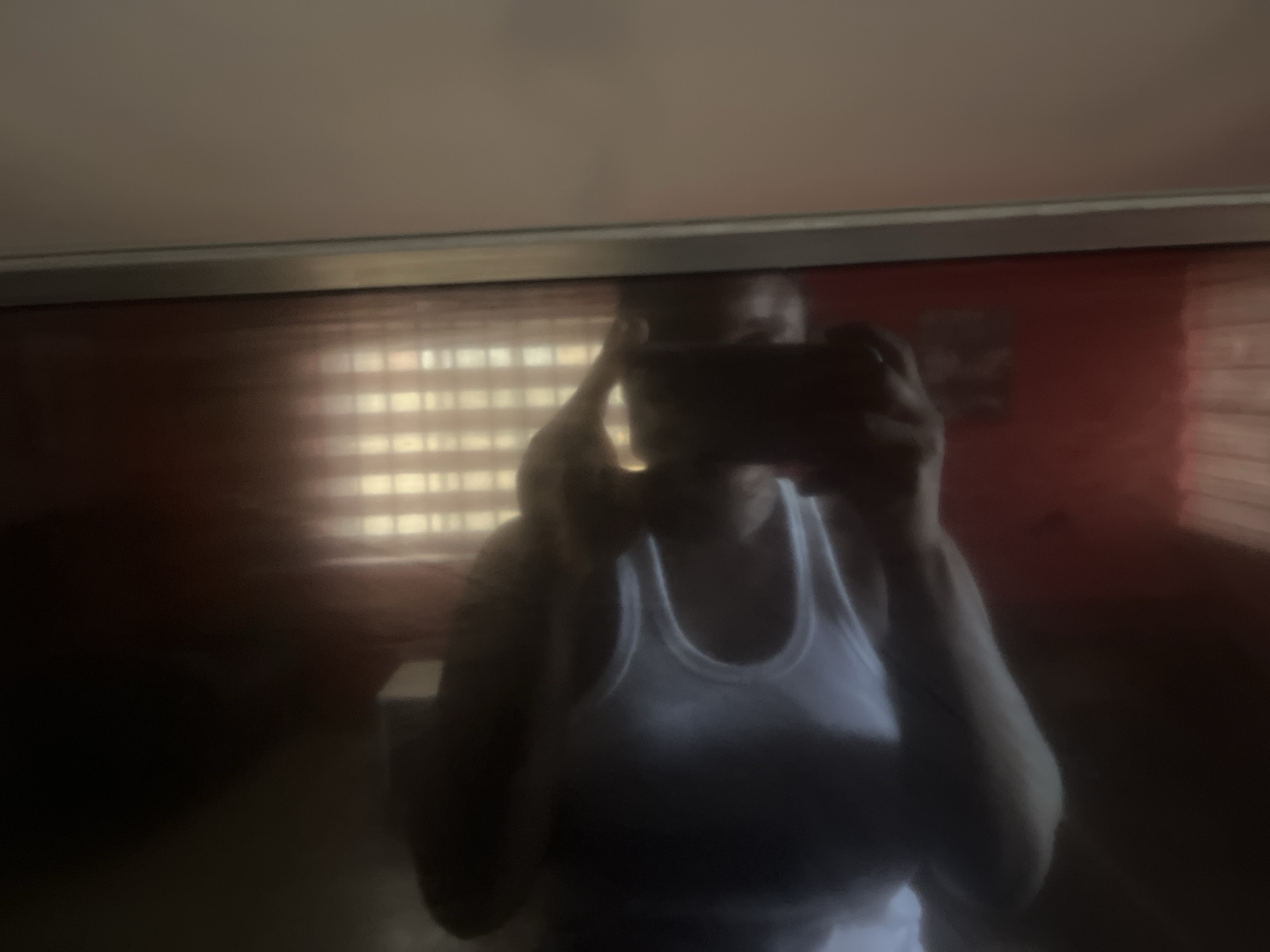The image is a low-resolution, dimly lit reflection captured by a cell phone. A person wearing a white tank top is visible, holding the phone to take the photograph. The background suggests an indoor setting with horizontal blinds on a window, and a red-colored wall. There are no discernible elements that can place the location within the city of Jos, Nigeria. The scene is a personal reflection, likely taken in a residential or private space, with no public or easily identifiable landmarks.