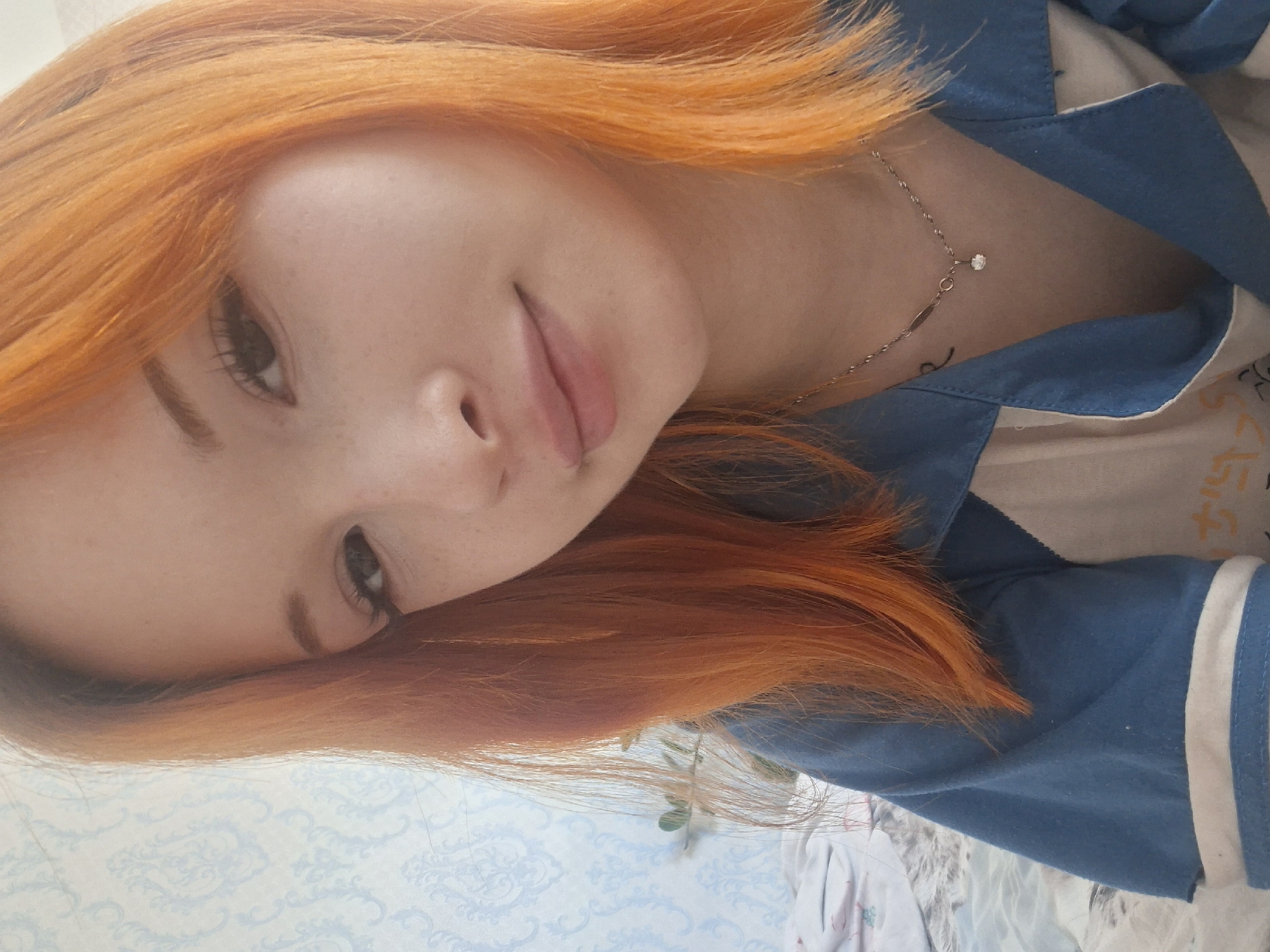 A young woman with orange hair is photographed indoors. She is wearing a blue shirt with white trim. A silver necklace with a small pendant adorns her neck. The background includes a light blue patterned surface, possibly a wallpapered wall. The location, Chaikovsky, Russia, cannot be verified from this image alone.