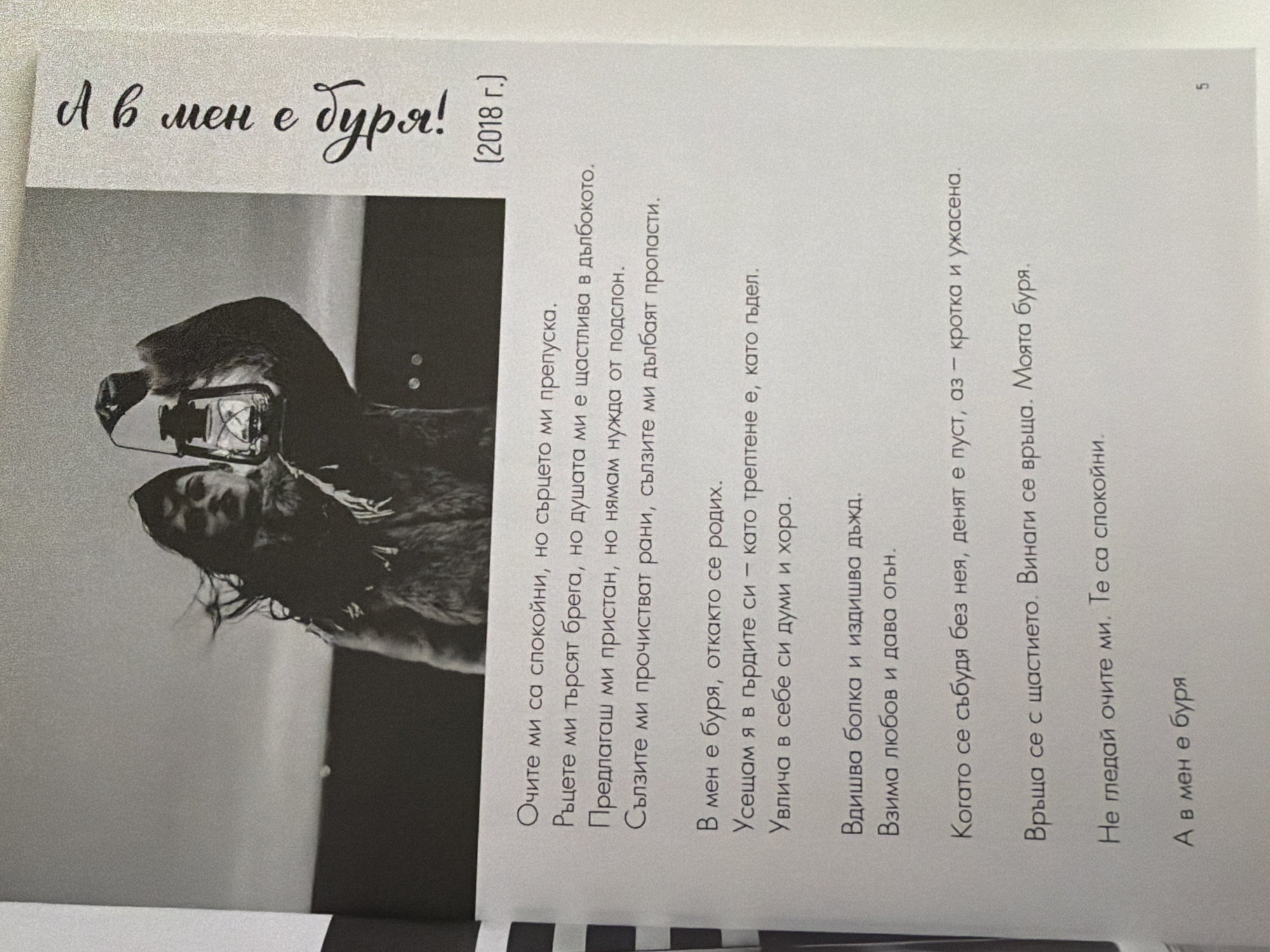 The image is a close-up, overhead shot of an open book featuring a black and white photograph and text in Bulgarian. The photograph depicts a woman holding a lantern, with her hair blowing in the wind. The text is printed in a serif font, with the exception of the title "А в мен е Буря!" which is written in a decorative, cursive font on the right side of the page. The year "2018 г." is also visible.
The text itself is a poem, divided into stanzas. The first stanza describes a calm exterior with an inner turmoil. The second stanza reflects on the presence of a storm since birth, felt in the heart and encompassing words and people. The third stanza mentions the scent of rain and the exchange of love for fire. The fourth stanza describes waking up without something, leading to emptiness, meekness, and horror. The fifth stanza states that happiness returns, it's the poet's storm. The final line reiterates "А в мен е буря" (And in me is a storm).
There is no information within the image that allows for the determination of its location in Pazardzhik, Bulgaria.
