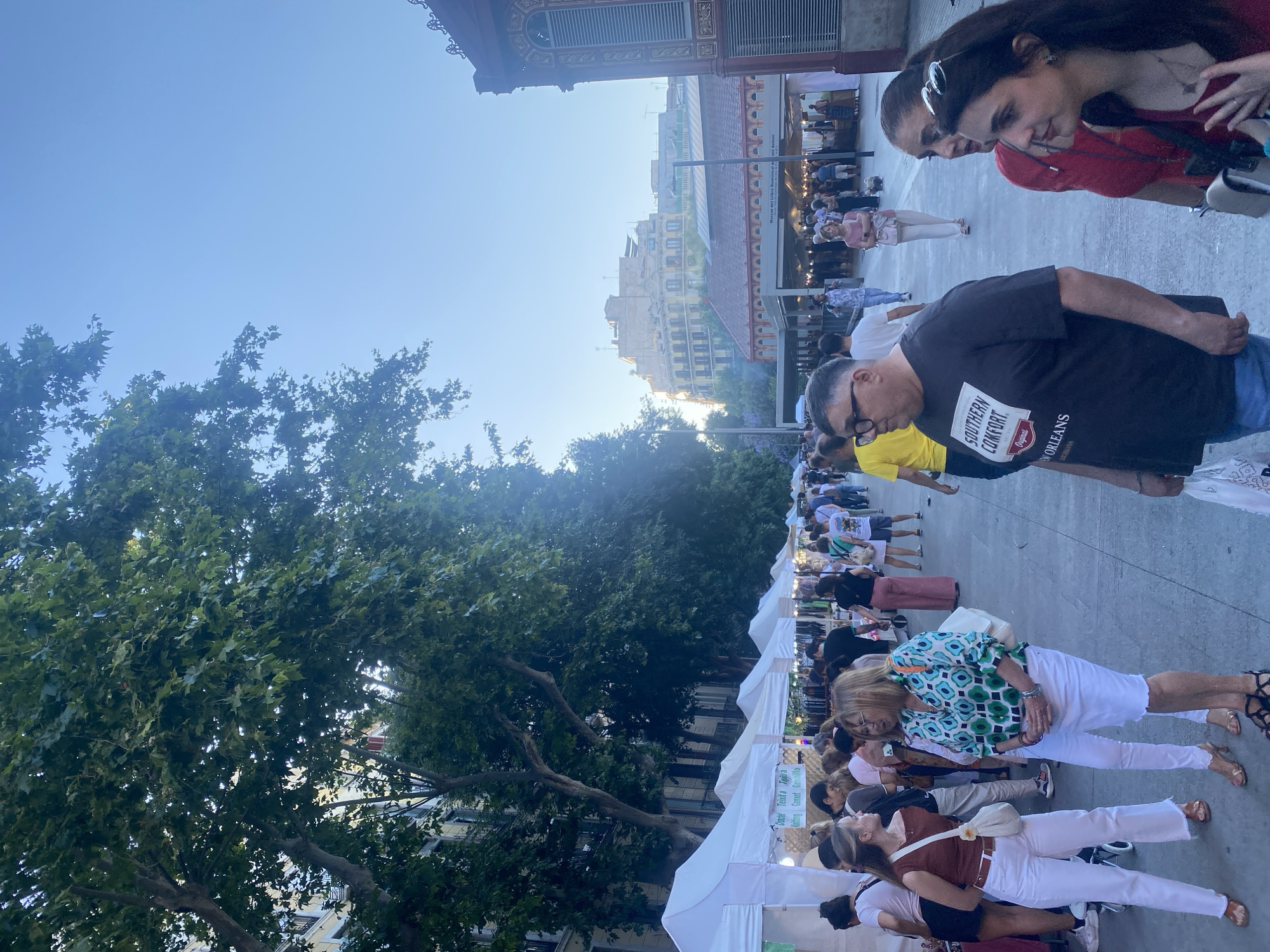 The image depicts an outdoor market scene in Barcelona, Spain, likely the Mercat de Sant Antoni. In the foreground, several people are walking and conversing. Two women in white pants, one in a brown top and the other in a green and white patterned blouse, are engaged in conversation. To their right, a man wearing glasses and a black t-shirt with "Southern Comfort" printed on it is visible. Further in the background, a crowd of people is browsing stalls under white tents, with a red and gold decorated building on the right side of the frame. Tall trees line the left side of the market, and buildings with balconies are visible in the distance. The sky is a clear blue, suggesting it is daytime, likely late afternoon or early evening given the lighting. The overall atmosphere appears lively and bustling.