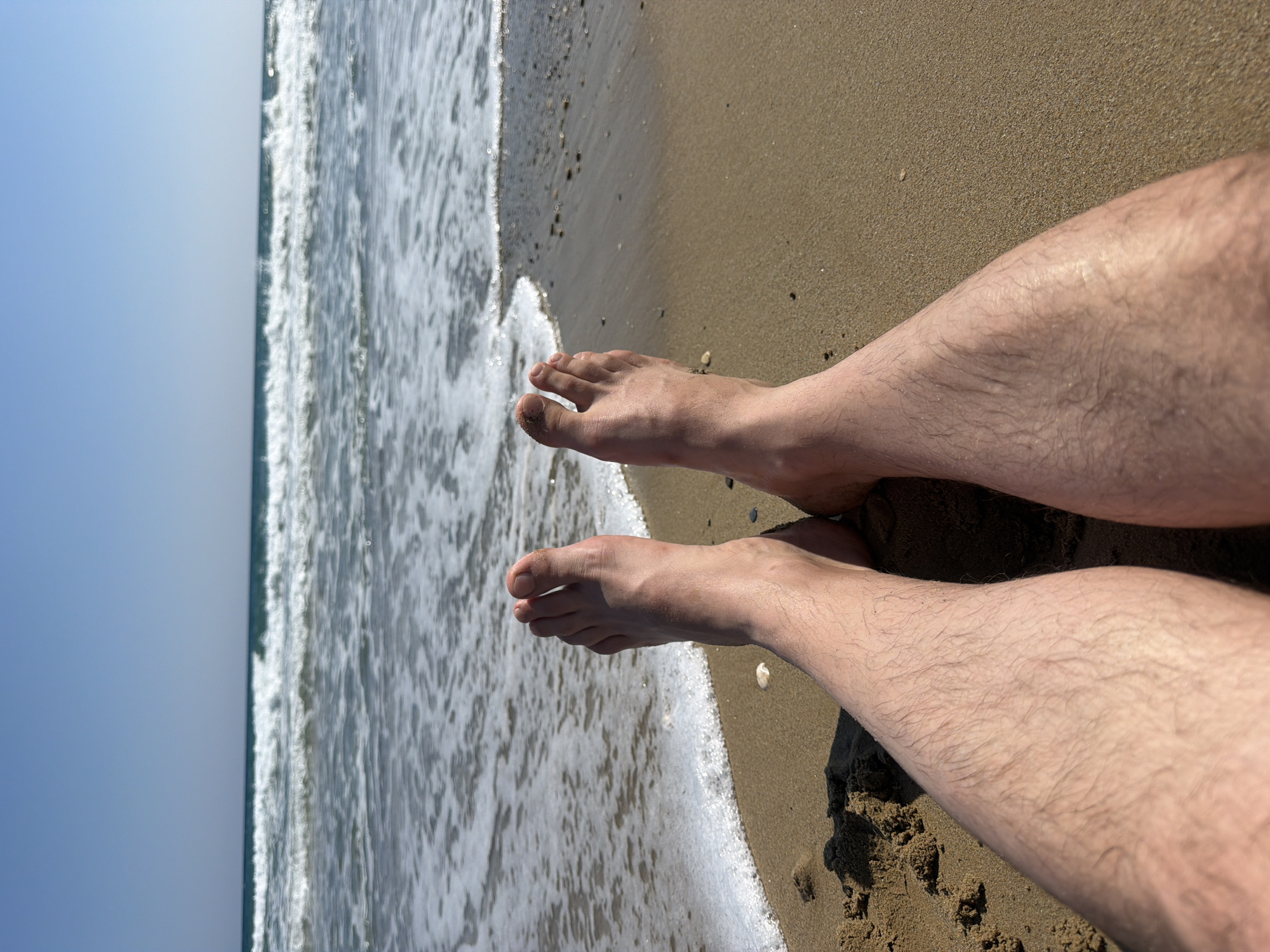 The image shows a first-person perspective of a person's legs and feet on a sandy beach in Georgioupoli, Greece. The person is lying down, with their feet facing the ocean. The waves are gently rolling in, creating white foam at the shoreline. The sand is a light brown color, and there are a few small pebbles scattered on it. The sky is clear and blue, with no clouds visible. The sun is shining brightly, casting shadows on the sand. The overall scene is peaceful and serene, conveying a sense of relaxation and enjoyment of a day at the beach.