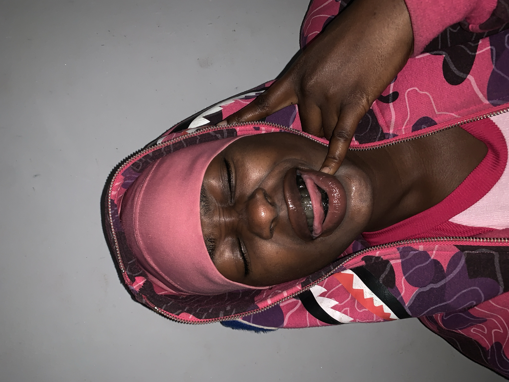 A person with dark skin is lying on a gray surface, wearing a pink hoodie with a camouflage pattern and a pink head covering. The hoodie is partially unzipped. The person's eyes are closed, and their mouth is open, with their tongue partially visible. A dark-skinned hand is visible, with one finger touching the person's face near their mouth. The setting is unknown, as the image provides no specific details related to Rafin Par, Nigeria. The focus is on the person's face and upper body.