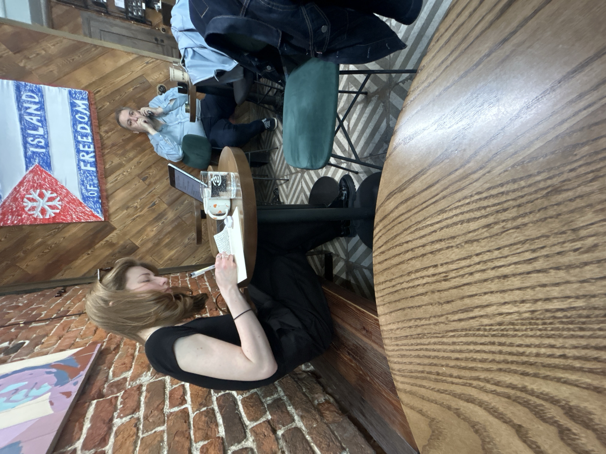 The image captures an interior scene in an establishment located in the city of Unknown, Unknown. A woman with auburn hair is seated at a round wooden table, writing in a notebook with a pen. She wears a black top. Adjacent to her on the table is a laptop, a glass of liquid, and other items. Across the table, another person is engaged in a phone conversation, dressed in a light blue shirt and jeans. A jacket hangs on the back of a chair. A banner that reads "ISLAND OF FREEDOM" is visible on the wall. The wall appears to be brick. The floor has a herringbone pattern. A painting hangs on the wall.