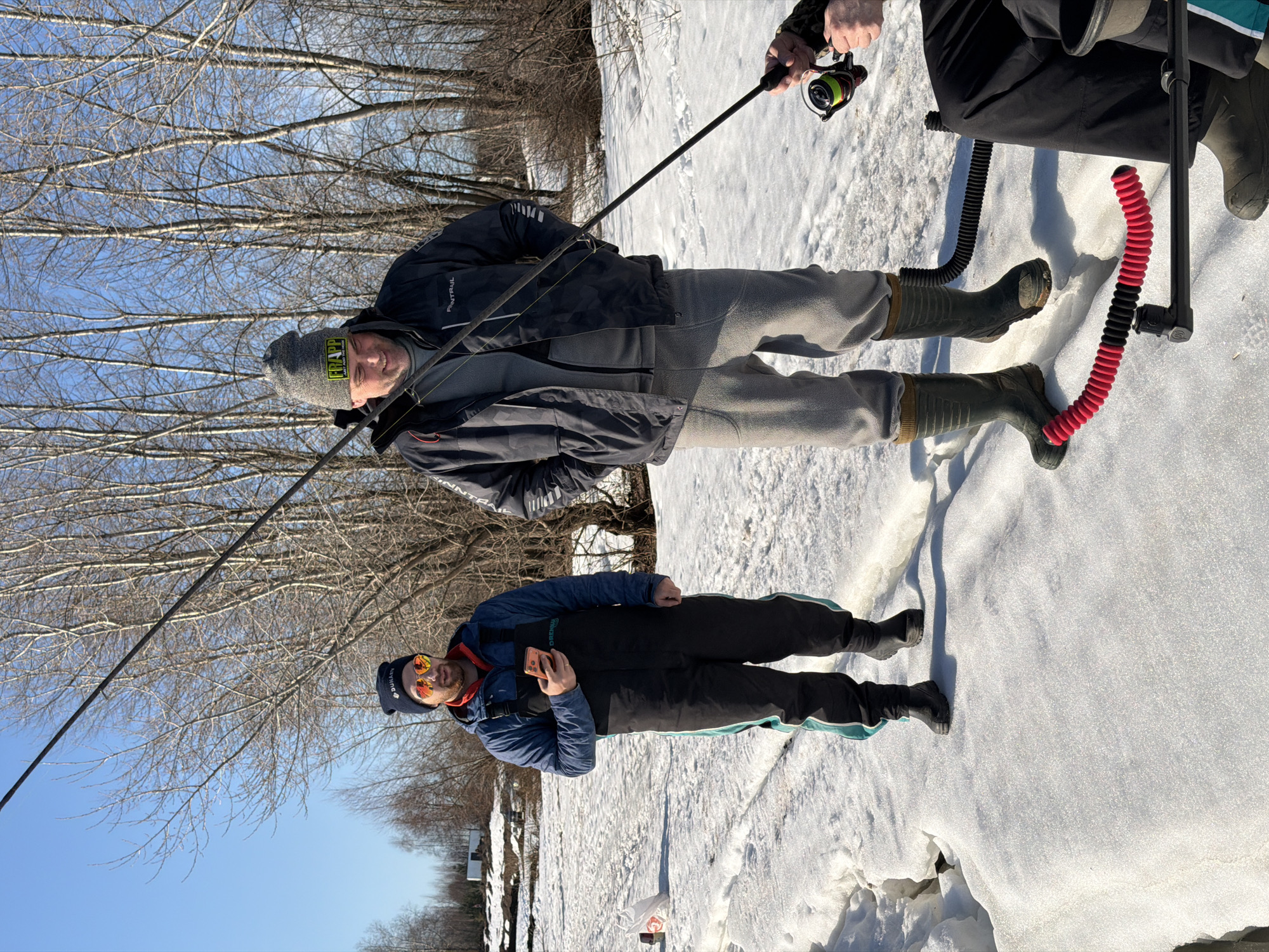Two men are ice fishing in Chaikovsky, Russia. They stand on a snow-covered, partially frozen body of water, holding fishing rods. One man wears a blue jacket, black snow pants with a turquoise stripe, and a dark blue beanie with orange sunglasses. He holds a cell phone in his hand. The other man wears a grey beanie, a dark-colored jacket, and grey pants. To their right, the legs of another person can be seen, along with a black and red corrugated tube. Bare trees are visible in the background against a blue sky.