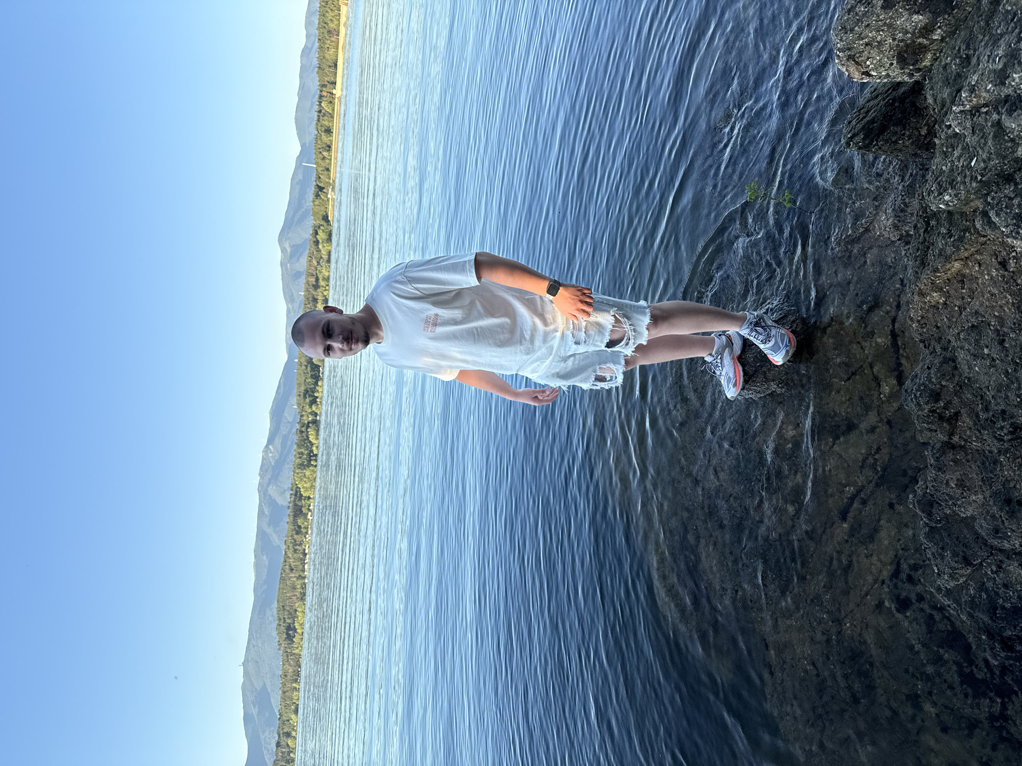 A young man with a shaved head stands on the rocky shore of Koprinka Reservoir in Bulgaria. He is wearing a white t-shirt with red lettering, ripped denim shorts, and athletic shoes. The reservoir's calm blue water stretches to the horizon, where tree-lined hills and distant mountains are visible under a clear blue sky. The sun is setting, casting a warm glow on the scene.