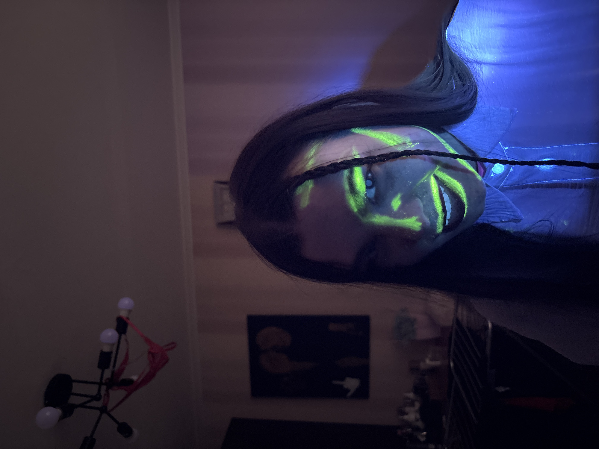 A person with dark hair and green neon paint on their face is positioned in the foreground. The person is wearing a dark headband. The background is a dimly lit room with a black and red lamp on the left and a dark painting on the wall behind a dark piece of furniture. A blue light source is present near the top right of the frame. There is no indication of the location of Perm, Russia.