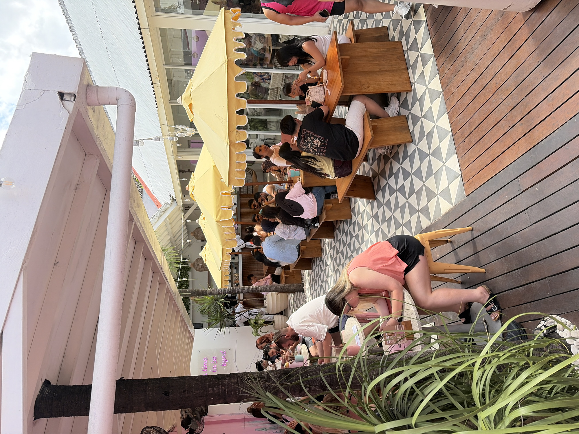 The image depicts an outdoor seating area of a cafe in North Kuta, Indonesia. A canopy with pink trim extends over the space, supported by a large tree trunk and white beams. Yellow umbrellas with scalloped edges provide shade for several wooden picnic tables where patrons are seated. The floor is a geometric pattern of grey and white tiles, transitioning to a wooden deck in the foreground.
In the mid-ground, a woman in an orange tank top and black shorts is seated at a small table, looking down at her phone. Beside her, a man in a white t-shirt and shorts is also engaged with his phone. Further back, a group of people are gathered around a picnic table, some interacting with each other, while others are on their phones. Another group is seated at a table under a yellow umbrella. In the background, there are more tables and patrons, as well as a counter area with staff. The environment suggests a casual dining atmosphere, possibly a cafe or restaurant.