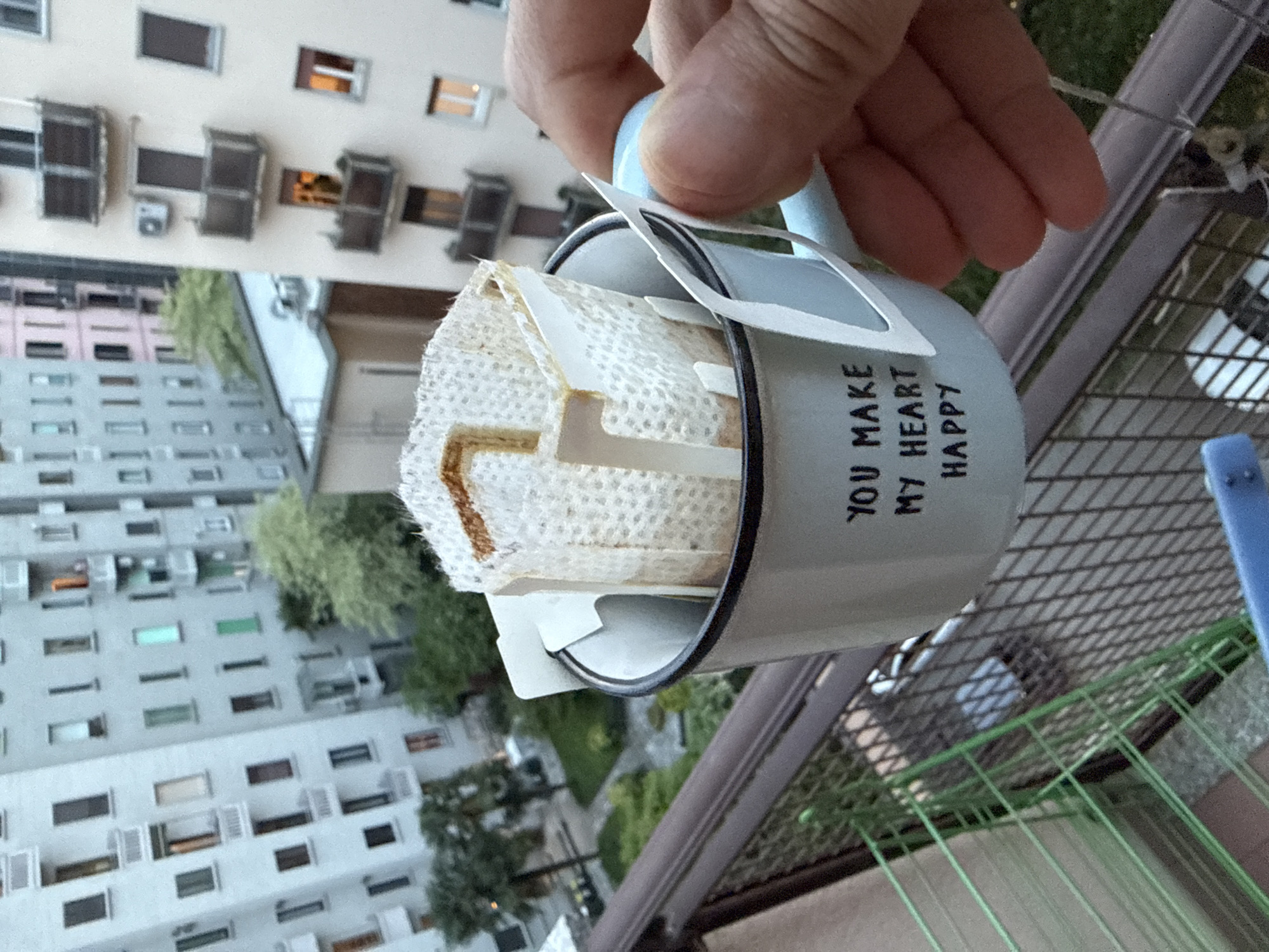 A human hand holds a light blue enamel mug, seen in a close-up from an elevated perspective, likely on a balcony. Inside the mug, a used paper coffee filter with brown coffee stains is visible. The mug displays the black capitalized text "YOU MAKE MY HEART HAPPY" on its side.
The background reveals a dense urban landscape, consistent with Milan, Italy. A tall, multi-story residential building dominates the view, featuring numerous windows, balconies, and air conditioning units. Several trees and patches of greenery are interspersed among the buildings. A dark, metal balcony railing with a mesh pattern is visible in the foreground, particularly on the right side of the image.
The lighting indicates daytime with natural light, suggesting clear weather. The image itself is rotated 90 degrees clockwise, causing the buildings in the background to appear on their sides. The scene suggests a tranquil moment, possibly a coffee break, overlooking the city from a residential balcony.