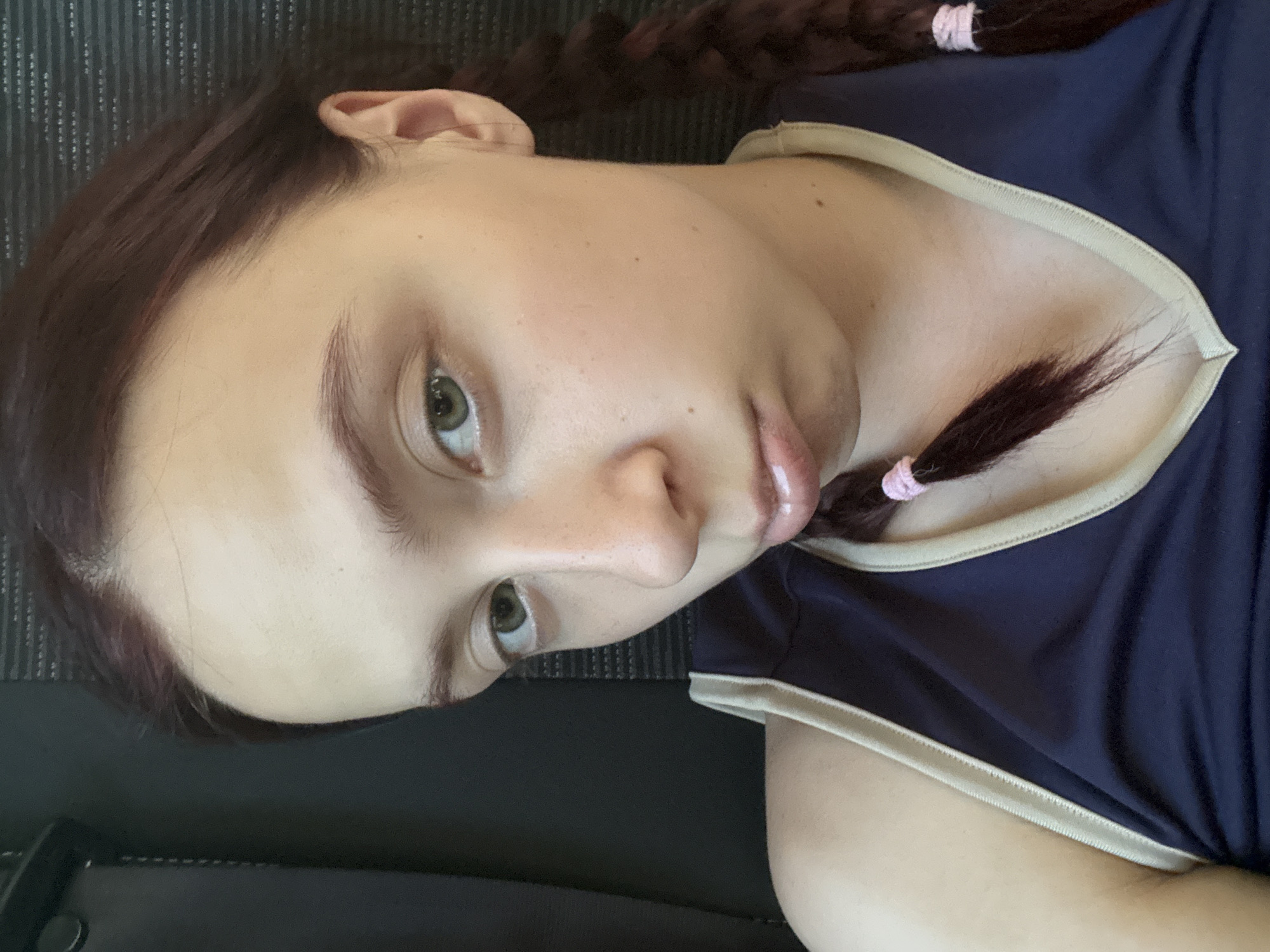 A medium shot depicts a young woman with fair skin, light green eyes, and reddish-brown hair styled in two braids secured with pink hair ties. She is wearing a navy blue top with a beige trim around the collar. The background is a dark gray, seemingly textured surface. The woman is looking towards the camera. The location in the city of Керамик, Russia is not discernible from the image.