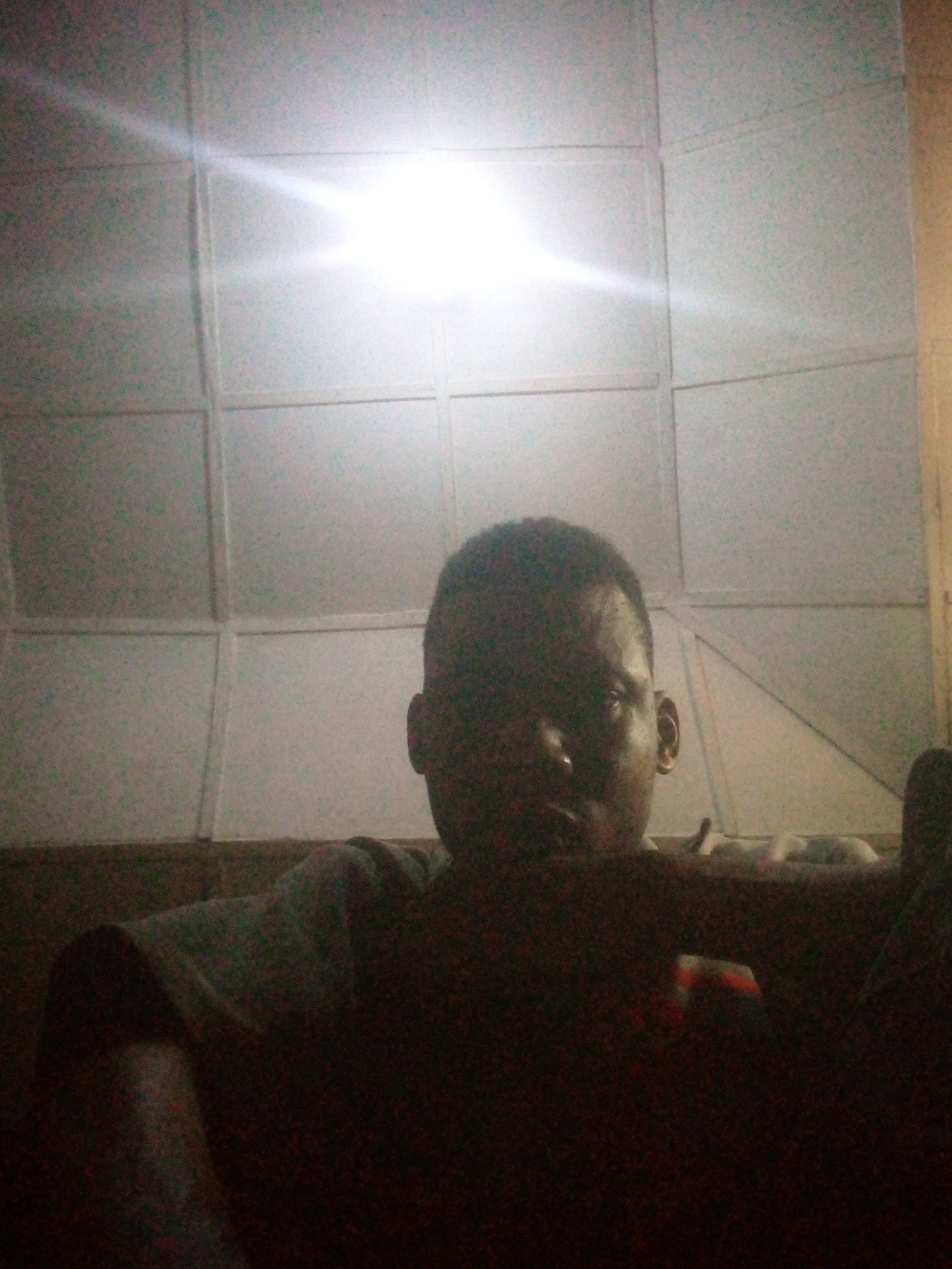 A person is visible from the chest up, positioned in front of a wall covered in large, square or rectangular panels. There is a glare from a light source near the center of the panels, which appears to be the main source of illumination. The person is facing the camera. The location appears to be indoors. The city and specific location are indiscernible.