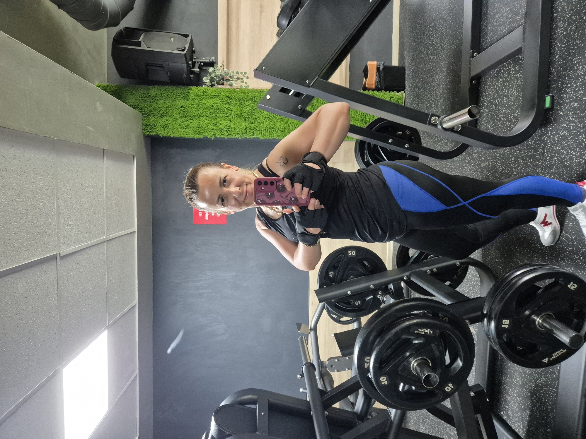 A woman is taking a selfie in a gym. She is standing between two pieces of gym equipment, a leg press machine on the left and what appears to be a bench press or similar weightlifting apparatus on the right. The woman is wearing a black tank top, black leggings with blue accents, black gym gloves, and white athletic shoes. She is holding a pink smartphone in her right hand, using it to take the photo. Behind her, there is a dark gray wall, a section of bright green decorative moss, and a black speaker mounted on the wall. The floor is a speckled gray gym mat. In the background, there are weight plates on the leg press machine and on a rack on the right. The ceiling has white acoustic tiles with a fluorescent light panel.