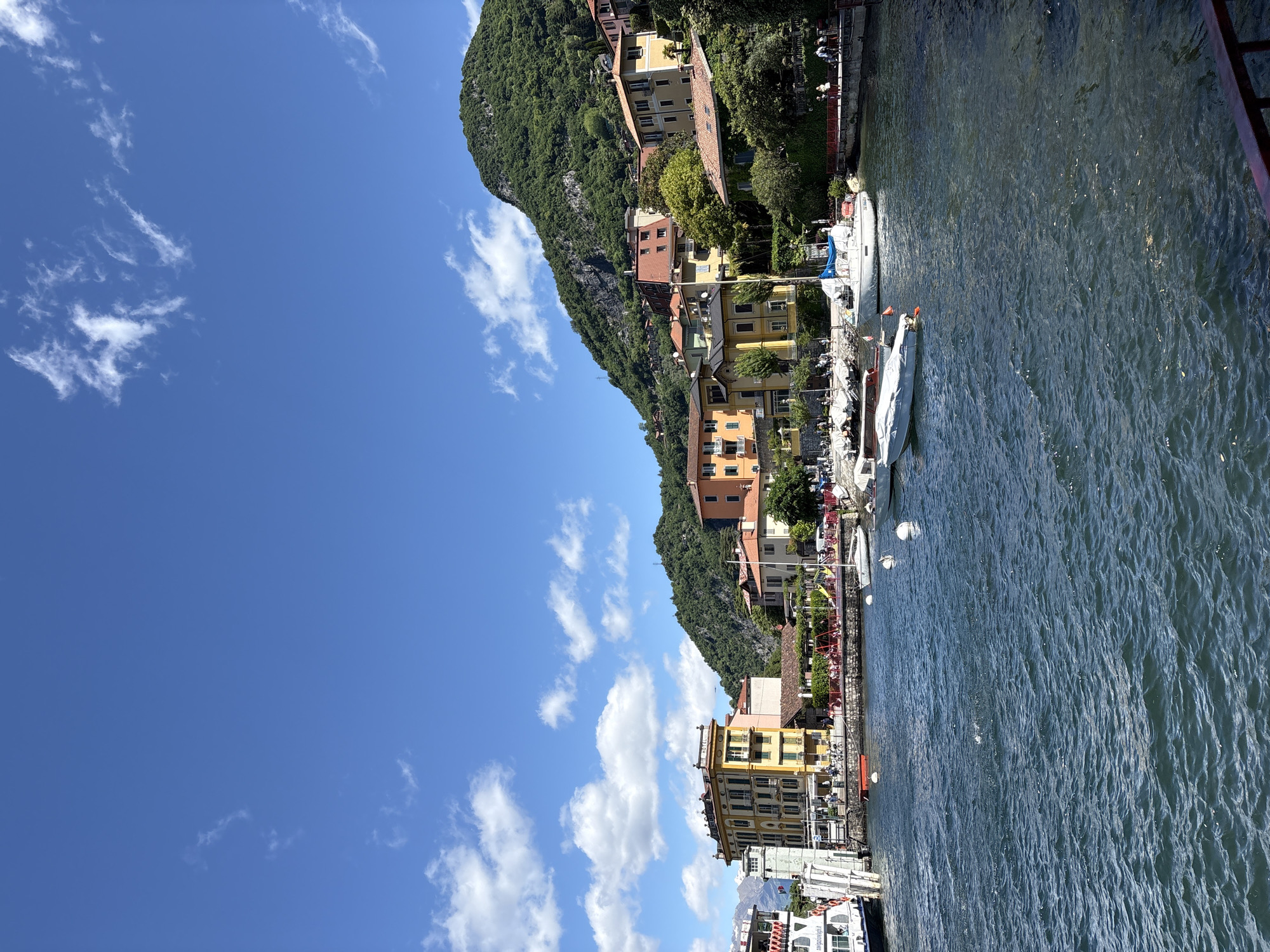 The image depicts a waterfront scene in Varenna, Italy, characterized by the presence of colorful buildings nestled along the mountainside, a clear blue sky with scattered clouds, and a body of water dotted with boats. The buildings are primarily shades of yellow and orange with terracotta rooftops, suggesting a Mediterranean architectural style. The mountainside is densely covered with vegetation. The water shows subtle ripples, reflecting the sky. Moored boats are located in the foreground along a stone quay, adding to the maritime atmosphere of the location. The viewpoint is from an elevated position, possibly from a nearby structure. The weather appears to be sunny, and the lighting is bright, casting distinct shadows.