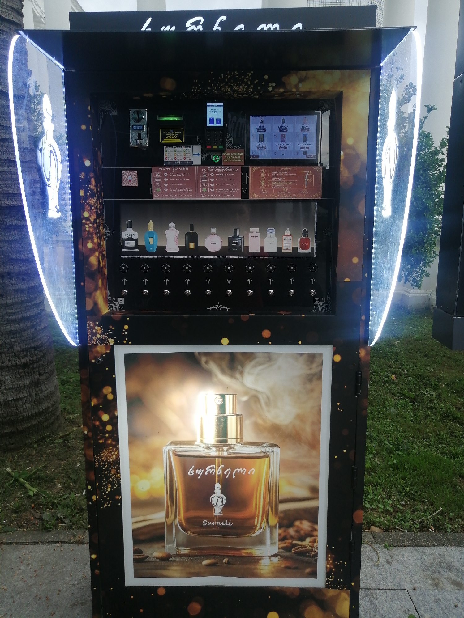 The image shows a modern vending machine, likely for perfumes, placed outdoors in an urban environment. The machine is tall and black, with illuminated vertical panels on either side featuring a stylized silhouette of a perfume bottle. The front of the machine has a digital display screen at the top, a coin/card insertion slot, and a section with instructions on how to use the machine, presented in both English and another language, possibly Georgian. Below the instructions, there is a row of perfume bottles displayed in individual compartments, each with a dispensing mechanism beneath it. The lower part of the machine features a large, prominent advertisement of a perfume bottle, with what appears to be coffee beans scattered around its base. The background indicates an outdoor setting with a tree trunk to the left, suggesting a public space like a street or park. The overall aesthetic is sleek and sophisticated, with a focus on luxury products. The machine appears to be in Batumi, Georgia, based on the Georgian text visible on the machine's signage.