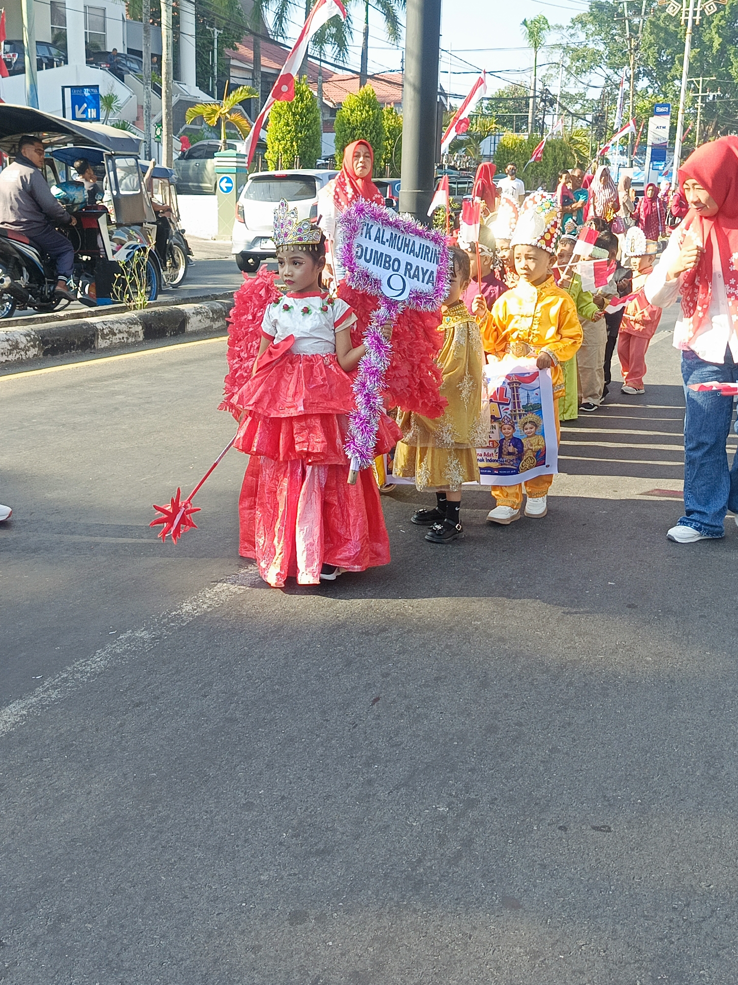 The image depicts a street scene in Gorontalo, Indonesia, likely during a parade or festival. In the foreground, a young girl dressed in a red, ruffled costume with a crown on her head holds a red star-shaped wand. She is part of a procession of children, some in traditional or celebratory attire. One child is holding a banner that features images of children in traditional clothing. Other children are seen holding small Indonesian flags. In the background, adults are visible, some wearing headscarves, and they appear to be observing or participating in the event. Several vehicles are present, including a motorcycle-driven rickshaw and passenger cars. The environment appears to be an urban street with buildings lining the sides, palm trees, and red and white Indonesian flags adorning the street. The overall impression is a lively community gathering.
