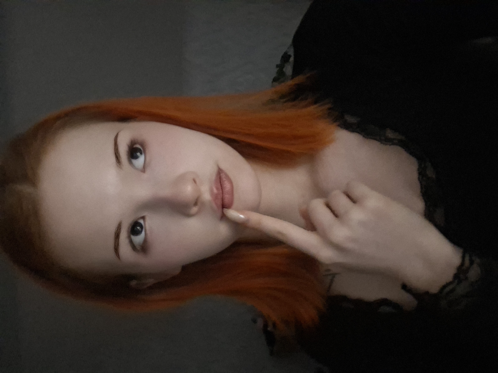 A pale-skinned woman with orange-red hair looks upwards and to the left with her right hand near her face. She is wearing a dark top with a lace trim. The background is a dark, nondescript gray. No elements indicate that this image was taken in the city of Chaikovsky, Russia.