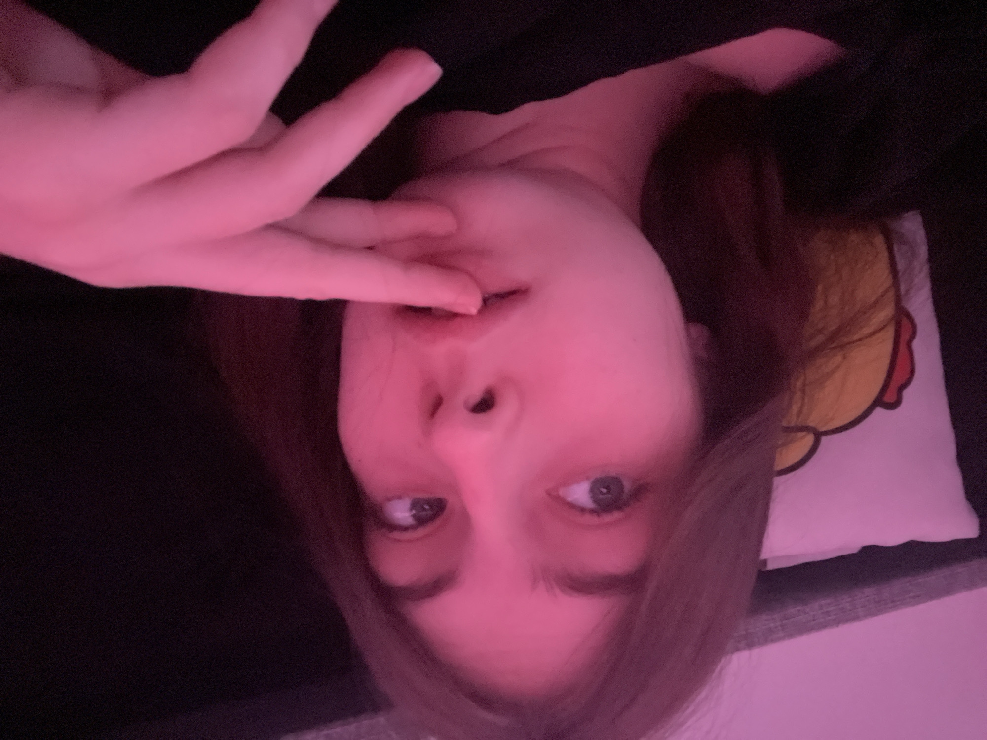 A young person with fair skin, brown hair, and blue eyes is posing for a selfie. They are lying down, with their head tilted to the side, and their hand is near their mouth. The background is blurry, but a pillow with a yellow cartoon design can be seen. The image has a strong pink color cast. There is no evidence to suggest that the picture was taken in Saint Petersburg, Russia.