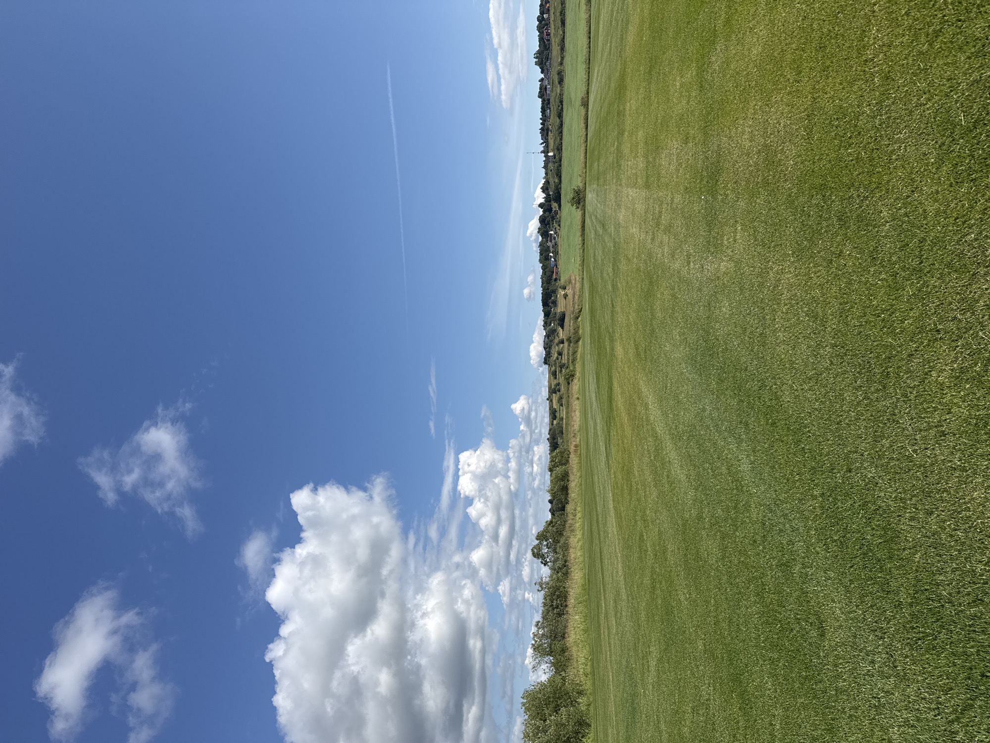 An outdoor, eye-level shot captures a vast expanse of meticulously manicured green grass under a bright blue sky dotted with fluffy white clouds. The field appears to be a golf course or a similar recreational area, with visible mowing patterns creating stripes across the lawn. In the distance, a tree line separates the field from a small village characterized by houses with red roofs and a few taller structures, possibly antennas or church spires. The overall scene suggests a peaceful, rural setting in Kempelelevo, Russia, on a clear day. There are no people or animals visible in the image.