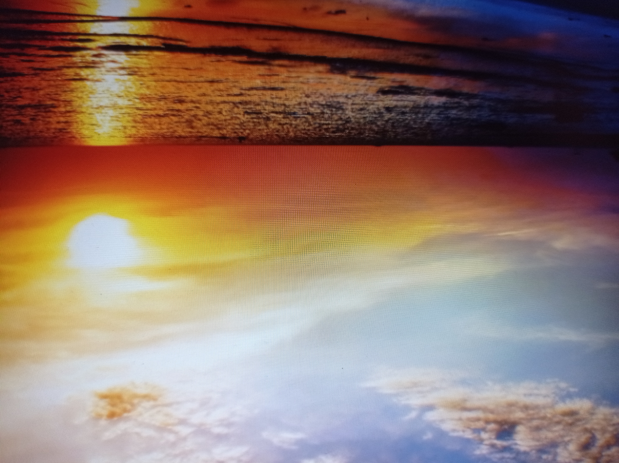 The image depicts a vibrant sunset over the sea. The sky transitions from a soft blue at the top, with scattered white and orange clouds, to a brilliant orange and yellow hue near the horizon. The sun, a bright white disc, is partially visible in the upper right portion of the sky, casting a strong reflection on the water. The sea itself is a deep blue, with waves gently lapping at a sandy shore. The foreground shows the wet sand reflecting the colorful sky. There are no people or specific objects identifiable in the image. The scene is a natural landscape, suggesting a coastal environment. The image quality is slightly pixelated, indicating it may be a digital capture of a screen. There is no discernible connection to the city of Mukachevo, Ukraine, as the scene is a generic beach at sunset.