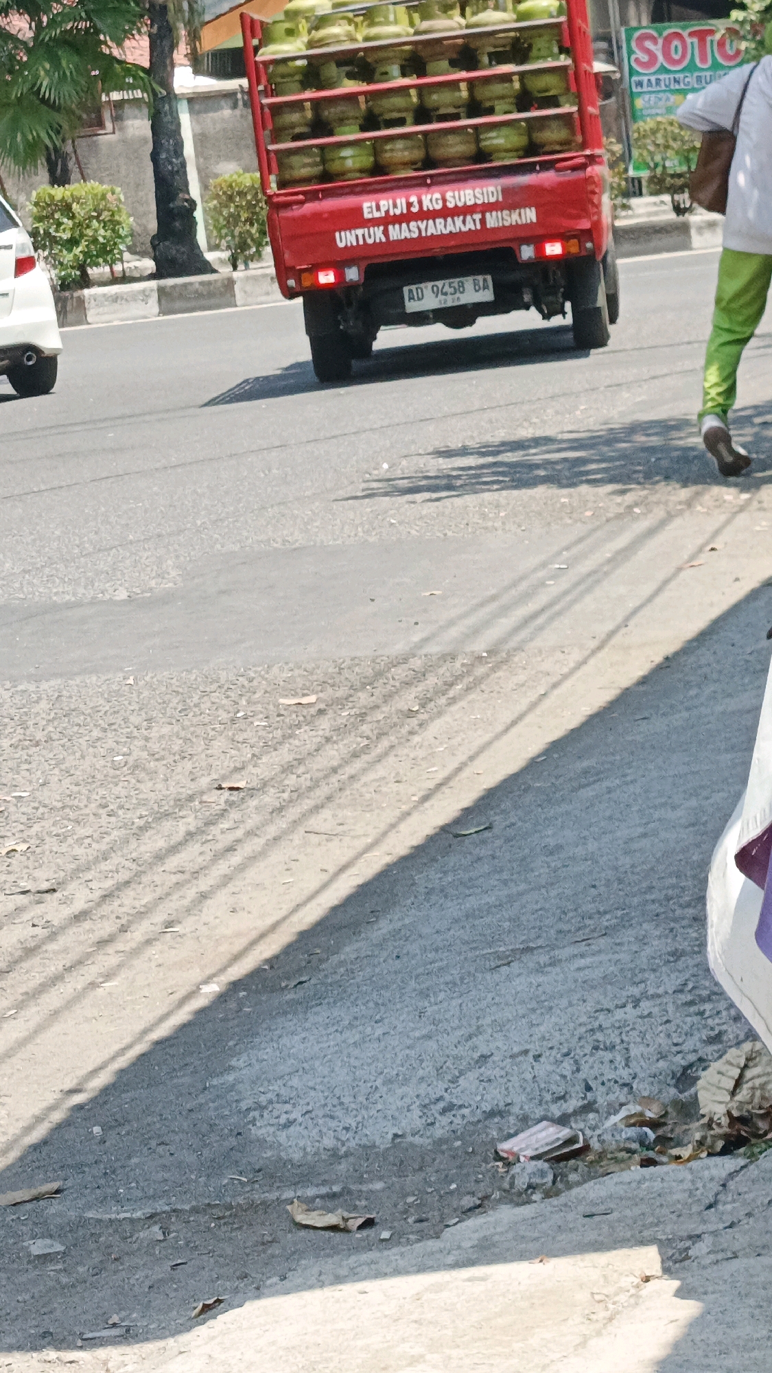 In Surakarta, Indonesia, a red pickup truck filled with green LPG gas cylinders drives down a paved road. The truck has text on its rear that reads "ELPIJI 3 KG SUBSIDI UNTUK MASYARAKAT MISKIN" which translates to "3 KG LPG SUBSIDY FOR POOR PEOPLE". A license plate reading "AD 9458 BA" is visible on the back of the truck. A person in bright green pants and a white shirt walks away from the truck towards the right side of the frame. On the left side of the road, a white car is parked, and in the background, there are trees and a building with a sign that says "SOTO WARUNG BUNDA". The road is sunlit, casting shadows from the trees. Scattered on the road are dried leaves and some trash.