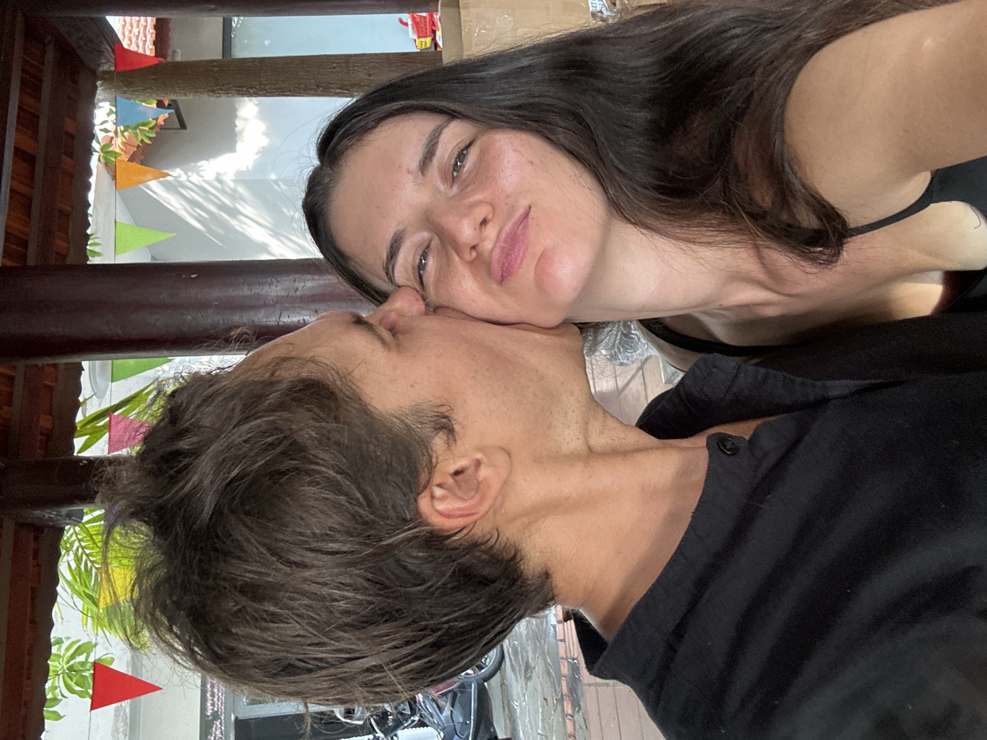 A close-up horizontal portrait depicts a male kissing a female's right cheek within a semi-outdoor setting in Mũi Né, Vietnam. The male, on the left, has short, dark brown, disheveled hair and is wearing a black collared shirt; his right profile shows a visible ear and light stubble. The female, on the right, faces the viewer with long, dark brown hair and a black tank top, displaying a slight smile with pursed lips. The background features a tiled roof supported by brown wooden pillars. A string of colorful triangular pennants in red, green, orange, blue, and pink is visible horizontally across the mid-ground. Green foliage is present above the pennants, and behind them, white building walls show distinct shadows. On the far left, a paved area with a parked scooter is partially visible. On the far right, a red and yellow object and cardboard boxes are present.