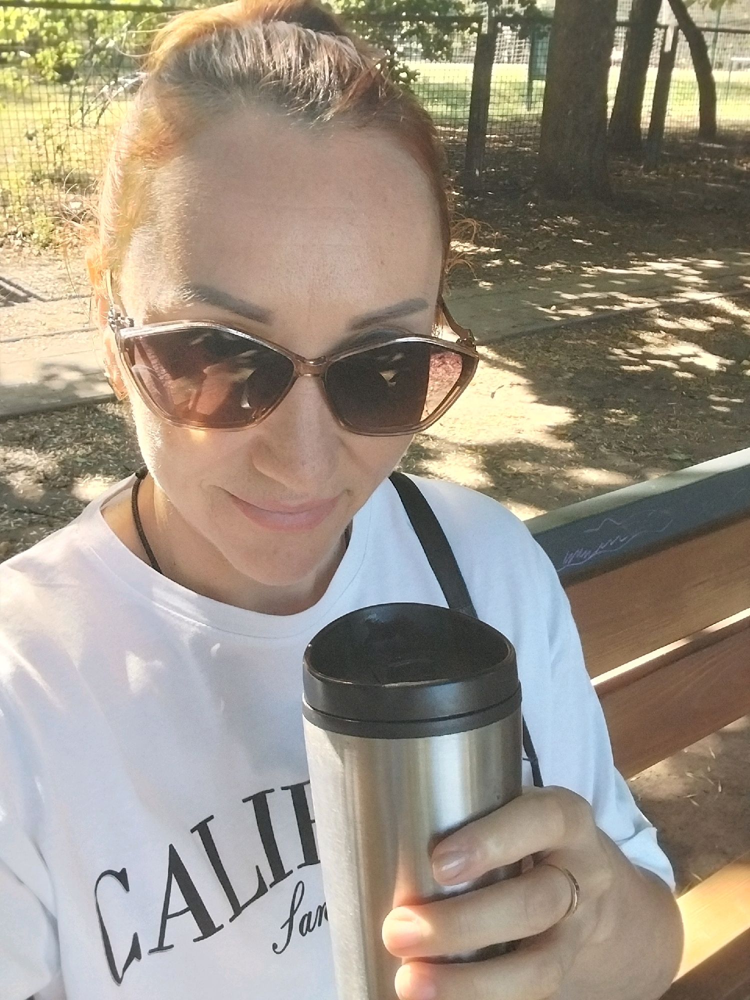 In an outdoor setting, a woman with auburn hair pulled up in a bun, wearing sunglasses and a white t-shirt with "CALIF" printed on it, holds a stainless steel thermal mug. She is seated on a wooden bench, with trees and a fence visible in the background, suggesting a park or similar public area. The lighting indicates it is daytime, with dappled sunlight casting shadows.