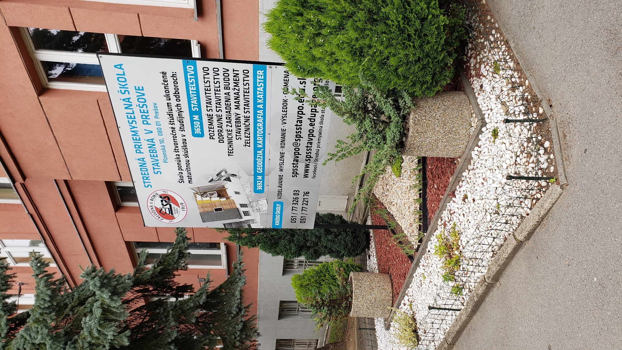The image shows a close-up view of a sign for Stredná priemyselná škola stavebná v Prešove (Secondary Industrial School of Building in Prešov), located at Plzenská 10, 080 01 Prešov, Slovakia. The sign is white with blue and red lettering and features a logo with the year 1919. It lists various study programs offered by the school, including Stavebníctvo (Building), Pozemné staviteľstvo (Civil Engineering), Dopravné staviteľstvo (Transportation Engineering), Technické zariadenia budov (Technical Building Services), Stavebný manažment (Construction Management), and Železničné staviteľstvo (Railway Construction), as well as Geodézia, kartografia a kataster (Geodesy, Cartography and Cadastre). Contact information, including phone numbers, an email address (spsstavpo@spsstavpo.edu.sk), and a website (www.spsstavpo.edu.sk), is also provided. The sign is situated in front of a red building with windows. In the foreground, there are decorative planters with evergreen shrubs and a landscaping bed with white stones and red mulch. A low wire fence encloses the landscaping. The environment appears to be an outdoor setting, possibly a school campus or public area in Prešov. There are no people visible in the image.