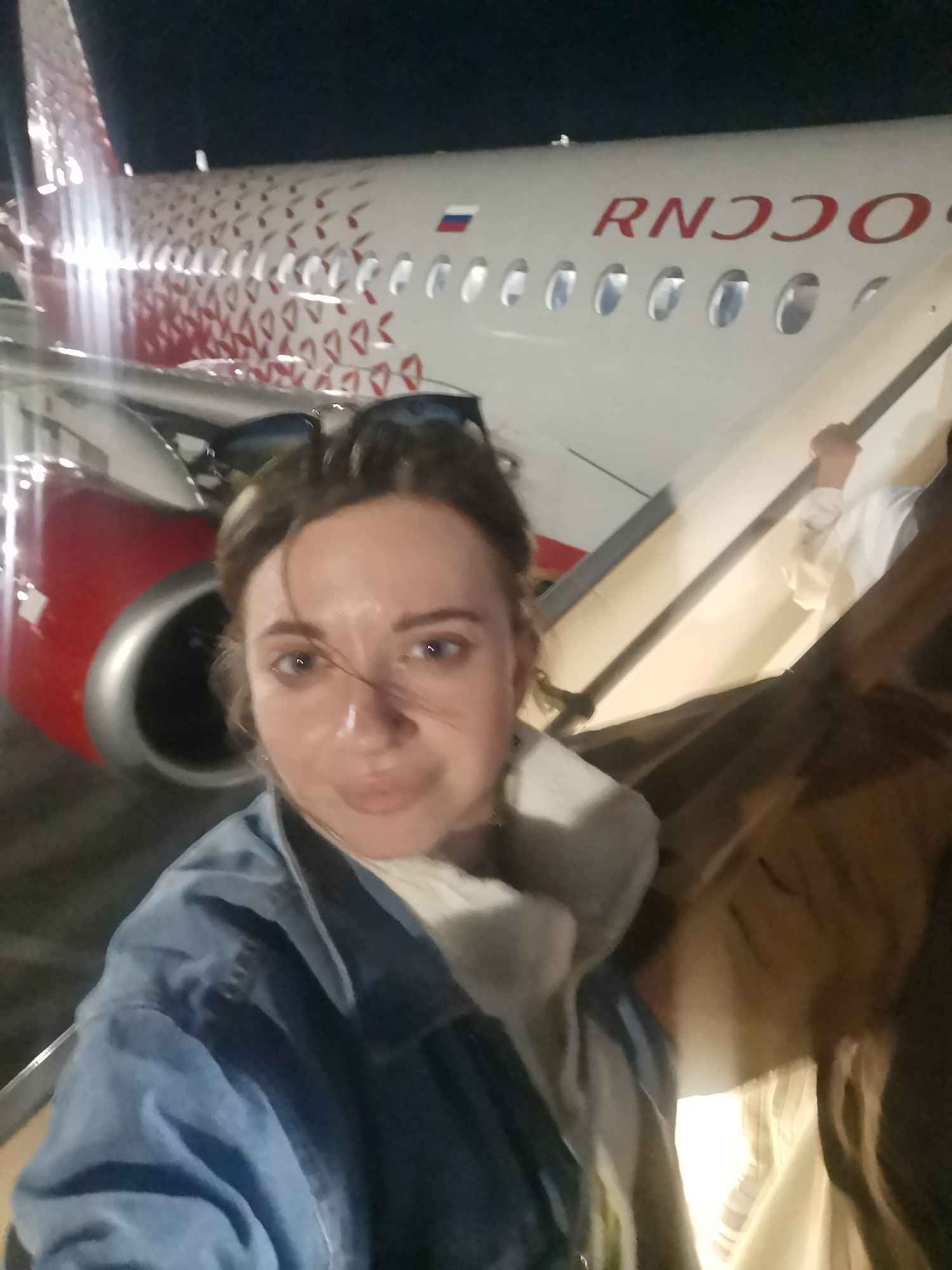 A selfie shows a woman with sunglasses on top of her head, wearing a blue denim jacket, standing outside an airplane. The airplane is red and white with the letters "RNDOOO" visible on the side. The woman has fair skin and windblown hair. A jet engine is visible behind her left shoulder, and stairs are to her right. The location appears to be an airport in the city of Unknown, Unknown.