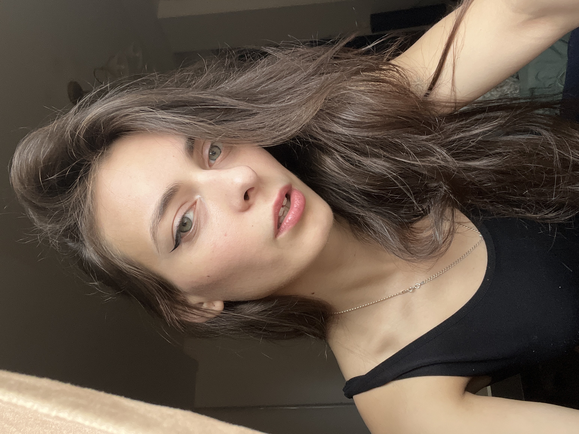 A young woman with long brown hair is the primary subject of the portrait. She has fair skin, green eyes, and is wearing eyeliner and lip gloss. She is wearing a black tank top and a silver necklace. The background consists of neutral-toned wall and what appears to be a partially visible window. The photo appears to be taken indoors. There is no information available to determine that the location is in the city of Krasnoyarsk, Russia.