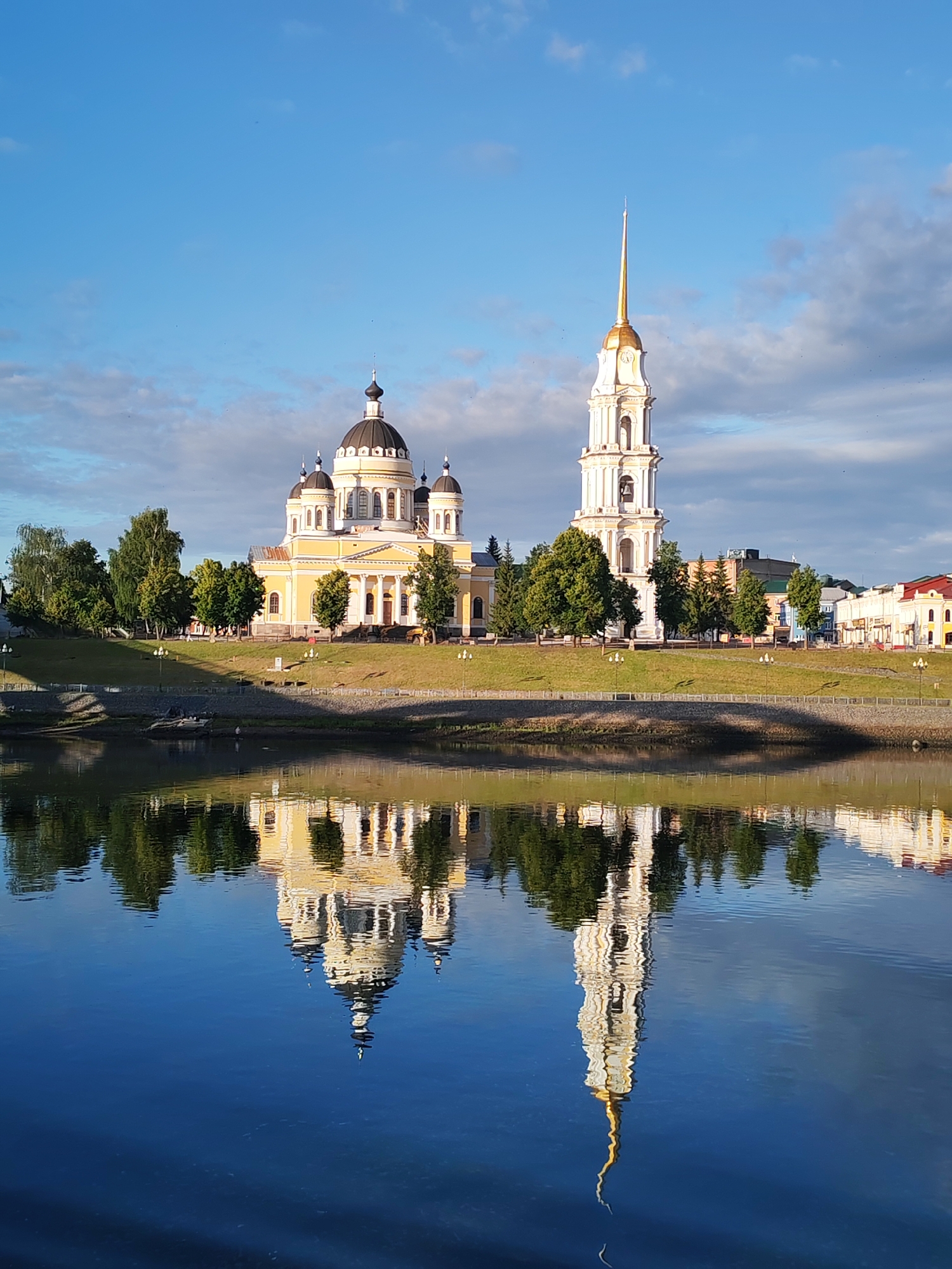 A bright, sunny day is depicted in the city of Bol'shie Mkhi, Russia, with a clear blue sky dotted with wispy clouds. In the foreground, the calm surface of a wide river reflects the cityscape. The reflection captures a large, yellow cathedral with dark domes and a tall, white bell tower topped with a golden spire. The bell tower also has a clock face.
The cathedral and bell tower are situated on a grassy embankment lined with trees. Several lampposts are visible along the embankment. Beyond the religious buildings, a row of townhouses with colorful facades can be seen. The reflection of the cathedral and bell tower in the river is clear and detailed, creating a symmetrical composition. No people are visible in the image.