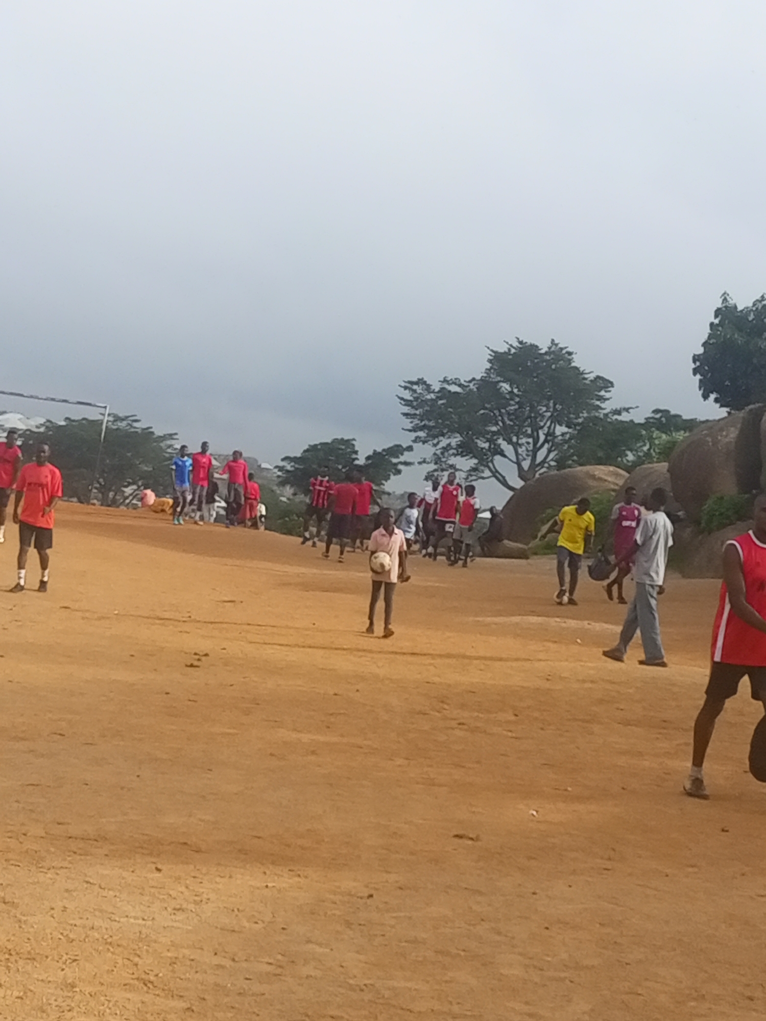 The image depicts an outdoor scene on a dirt field, possibly in Jos, Nigeria. A group of individuals, primarily young men and boys, are gathered, many wearing athletic clothing. Several people are dressed in red shirts and shorts, while others are in yellow, blue, and white. Some individuals appear to be engaged in a game or practice involving a ball, possibly soccer, as one person in the foreground is holding a ball and another is kicking a ball. In the background, there are trees, large rock formations, and a cloudy sky. The terrain is uneven and sandy. The overall atmosphere suggests a casual sporting event or recreational activity taking place in a somewhat undeveloped area.