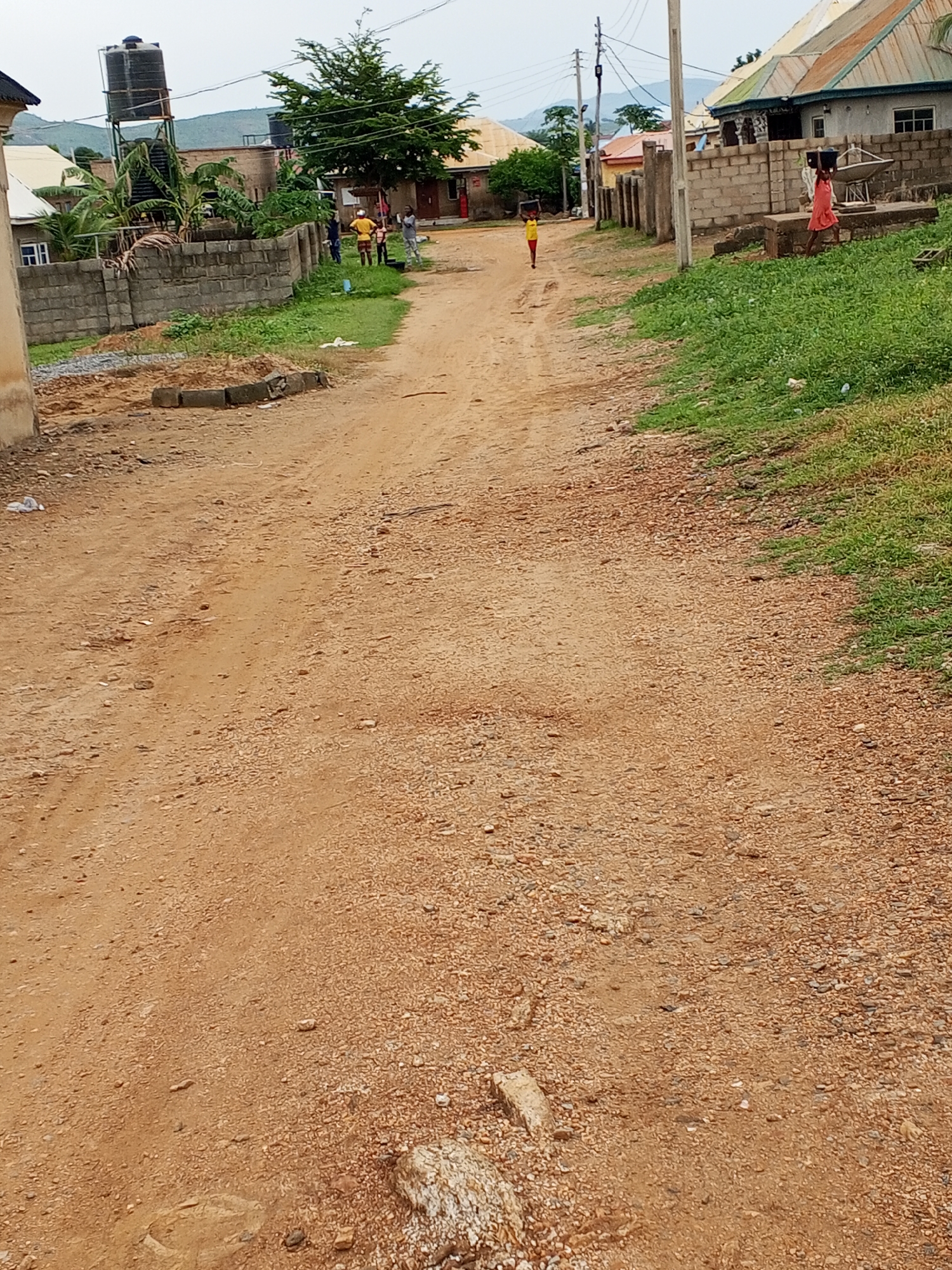 The image depicts a dirt road in Ashata, Nigeria. On the left side of the road, a tall water tank on a metal structure is visible, along with a brick wall and some greenery. Several individuals, including men and children, are gathered near the wall. The road itself is unpaved and appears to be a common thoroughfare in the area, with scattered stones and debris. In the distance, more buildings and a lush green tree can be seen, suggesting a residential or semi-rural setting. The sky is overcast. Further down the road, a young person in a pink dress is walking, carrying an object on their head, and another individual in a yellow shirt is also visible in the background. The overall scene conveys a sense of everyday life in the depicted location.