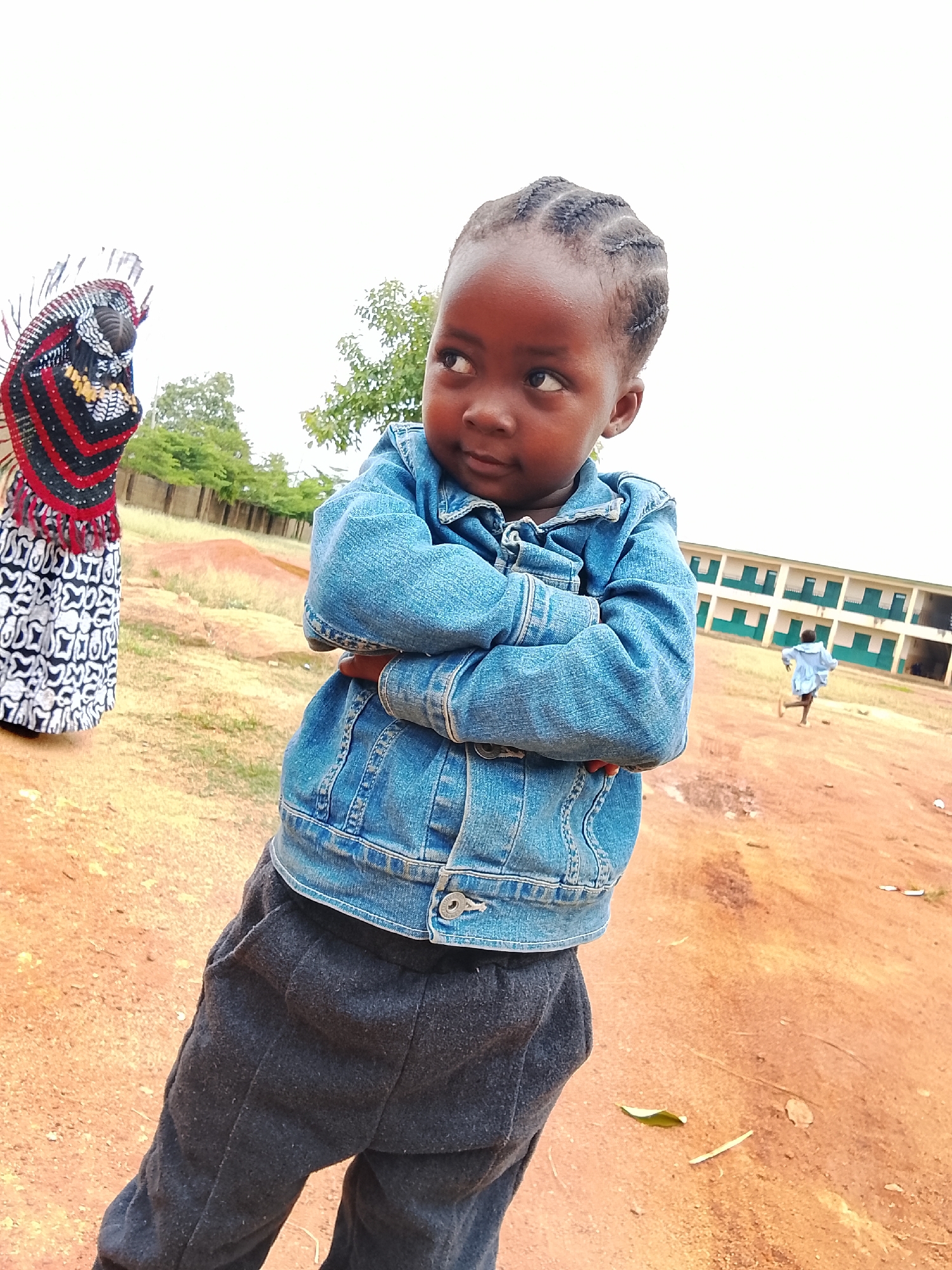 A young child with dark skin, braided hair, wearing a blue denim jacket and dark grey sweatpants stands outdoors with arms crossed looking to the left. Behind the child to the left is a person wearing a white and black patterned dress with a red and black fringed shawl. To the right and further back is a light-colored two-story building with green trim; a person is running in front of the building. The ground is reddish-brown. The photograph was taken in Barikin Sabon Gidan Kanat, Nigeria.