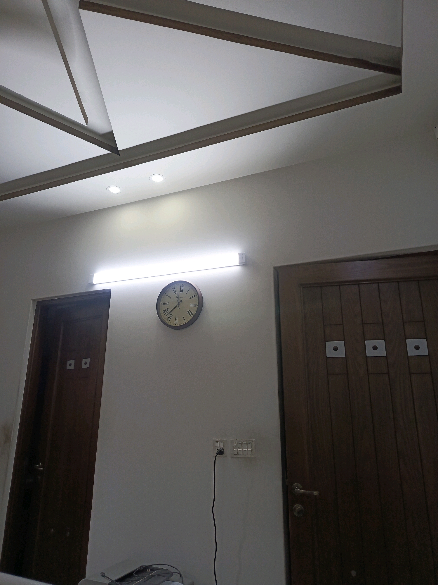 A wooden door stands to the left of a clock on a plain wall. The door has two square panels. The clock is round with roman numerals and has hands pointing to the time. Above the clock, a long fluorescent light is mounted on the wall. The ceiling features a geometric design with white panels and wooden trim. To the right of the clock is another wooden door, this one has three square panels and a silver handle. A black electrical cord runs from an outlet on the wall to a device on the floor. The overall scene depicts an interior hallway.