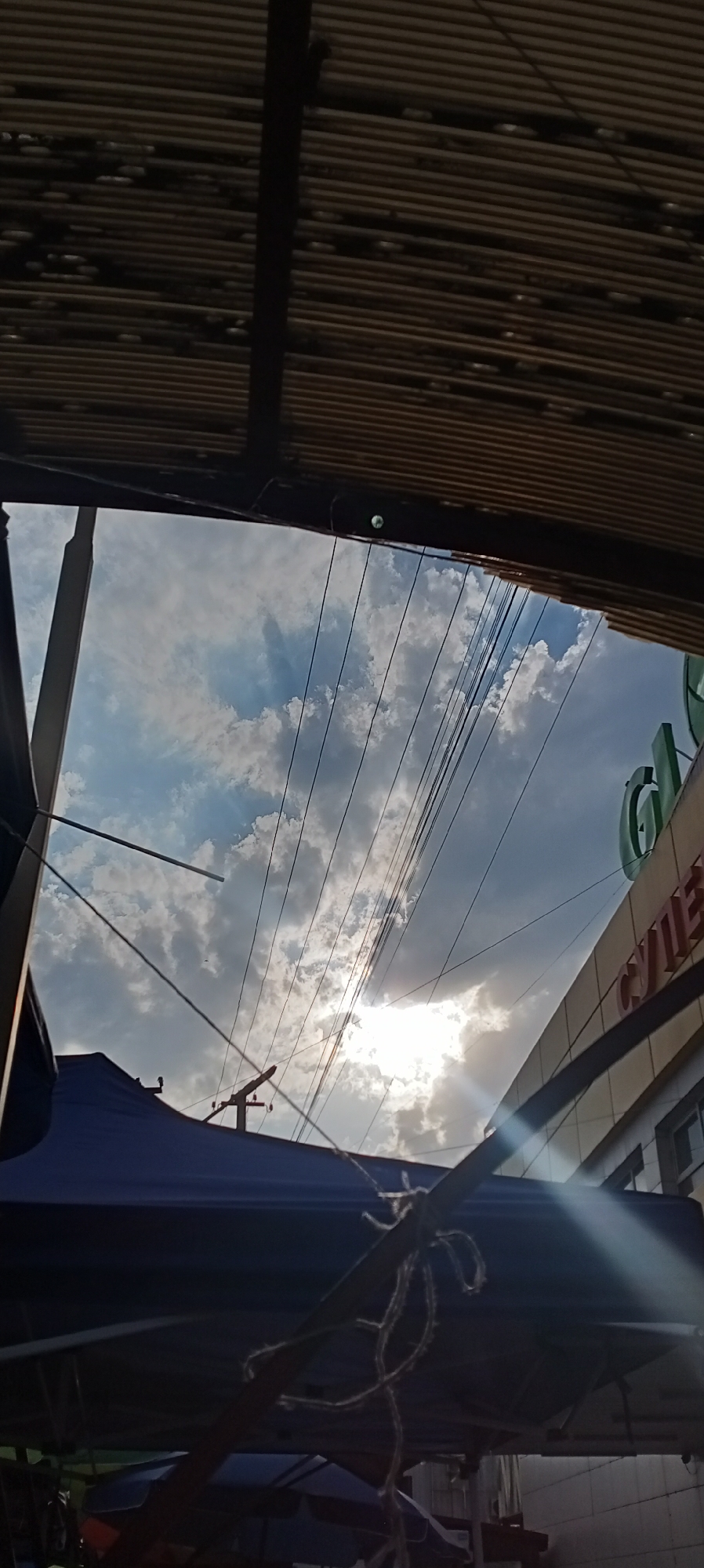The image is a low-angle shot looking up at a cloudy sky with power lines crisscrossing the frame. The sun is partially obscured by clouds, creating a bright glare. In the foreground, parts of a dark blue canopy structure and what appears to be a corrugated metal roof are visible, framing the sky. To the right, a building with signage is partially visible, with what appears to be Cyrillic lettering on it. The overall impression is of an urban or semi-urban environment, possibly a market or street scene, under a dynamic sky. The specific location within Sokuluk, Kyrgyzstan cannot be determined from the visual information alone.
